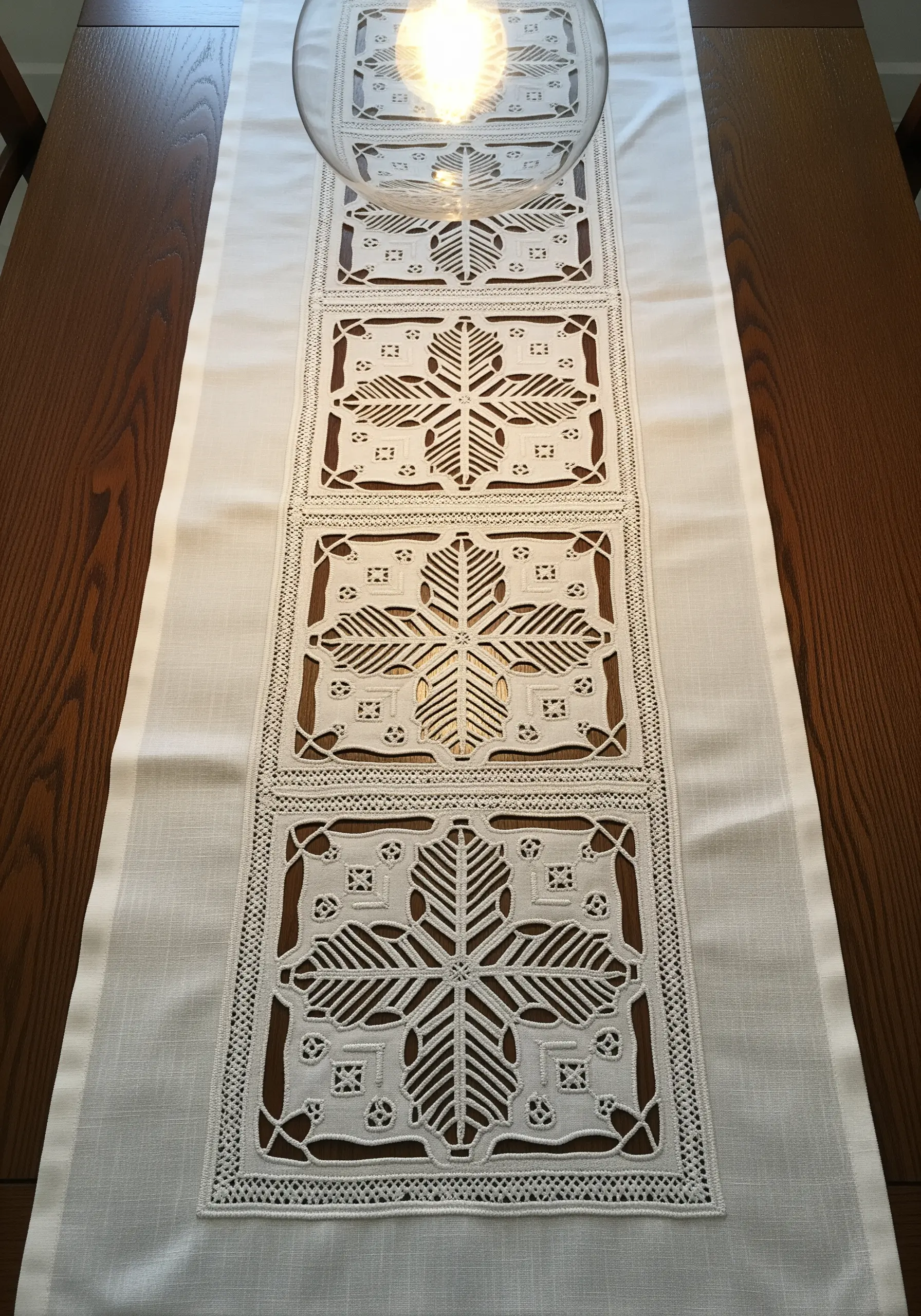 A white linen table runner with intricate cutwork embroidery creating geometric, lace-like patterns.
