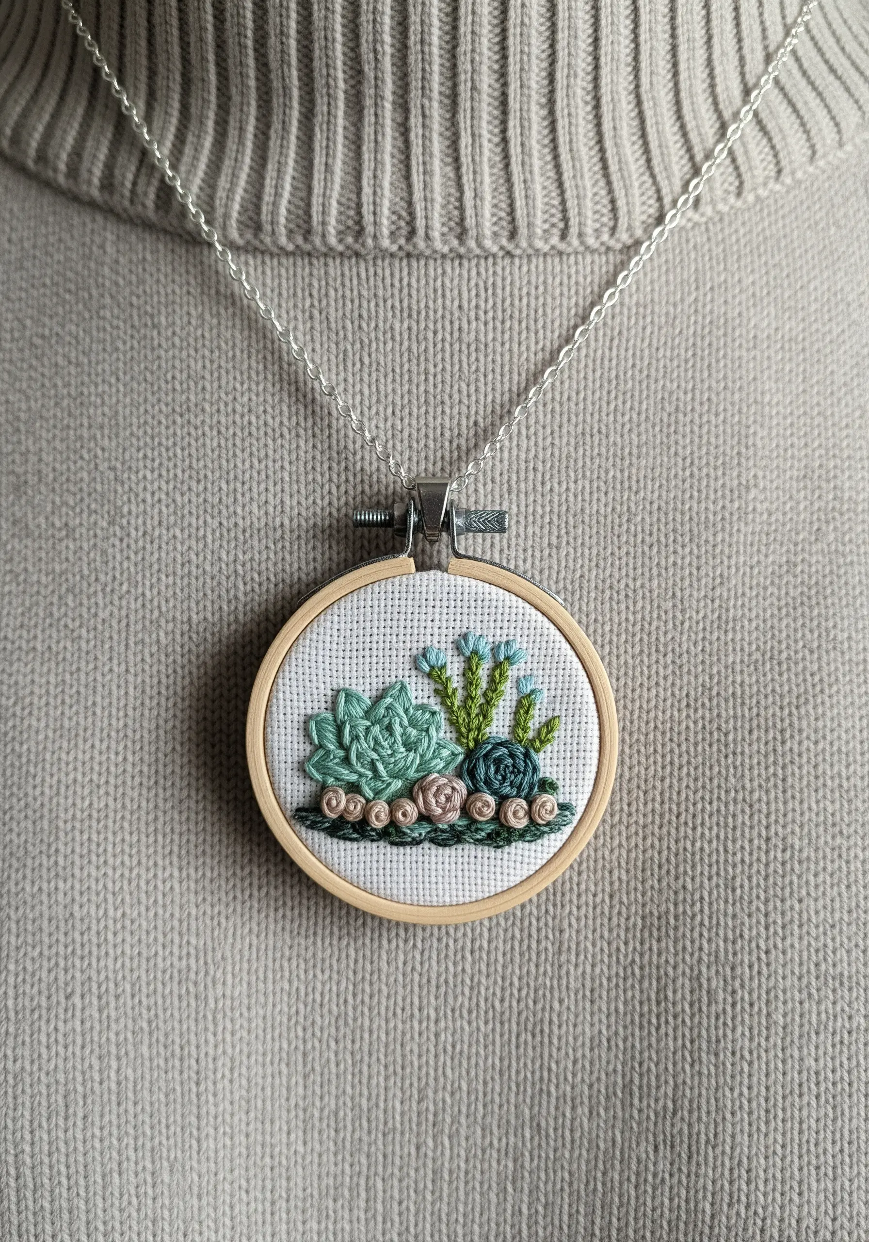 A small embroidery hoop necklace featuring a detailed, textured succulent garden.