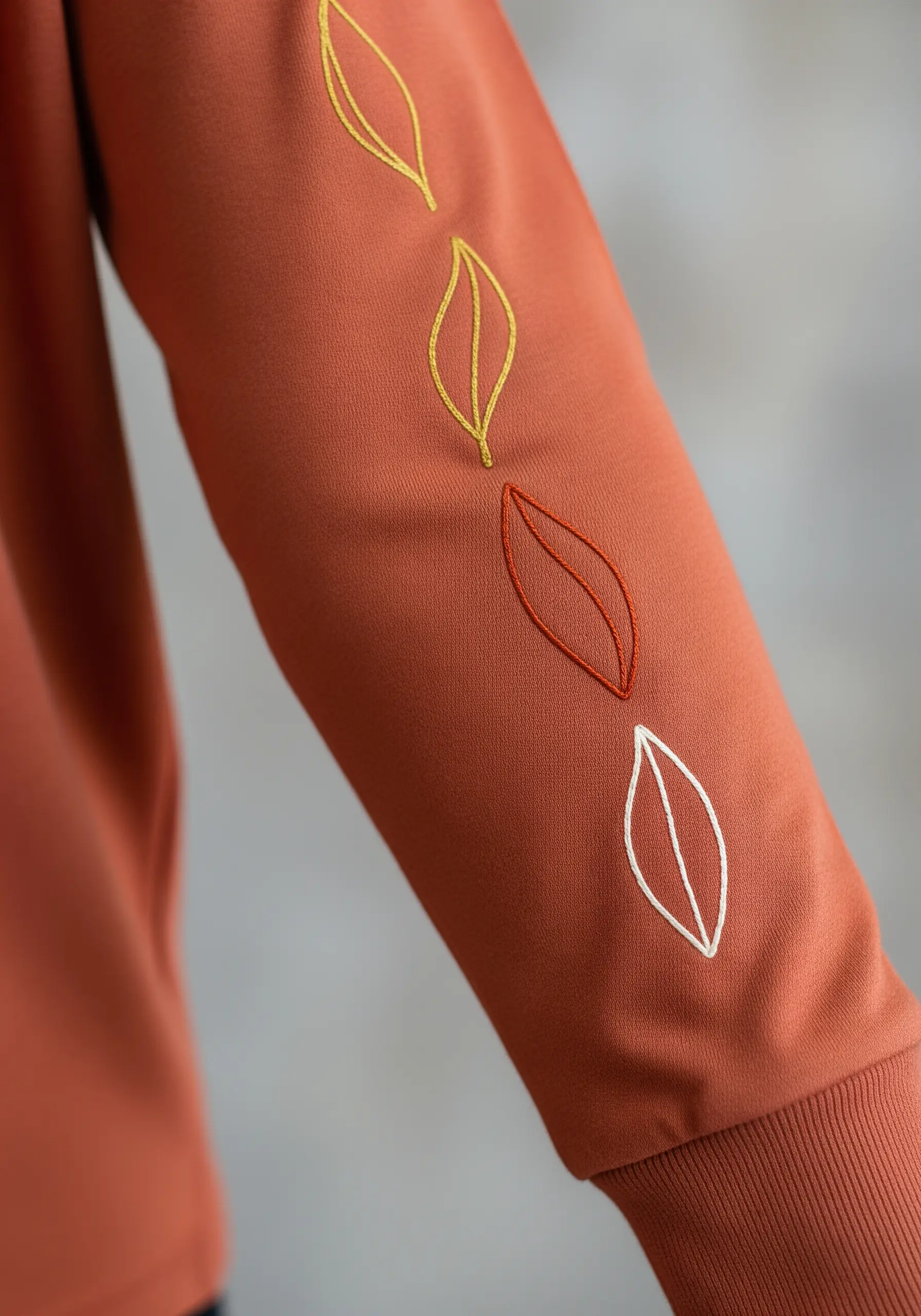 A series of leaf outlines in autumn colors stitched down the sleeve of a rust-colored shirt.