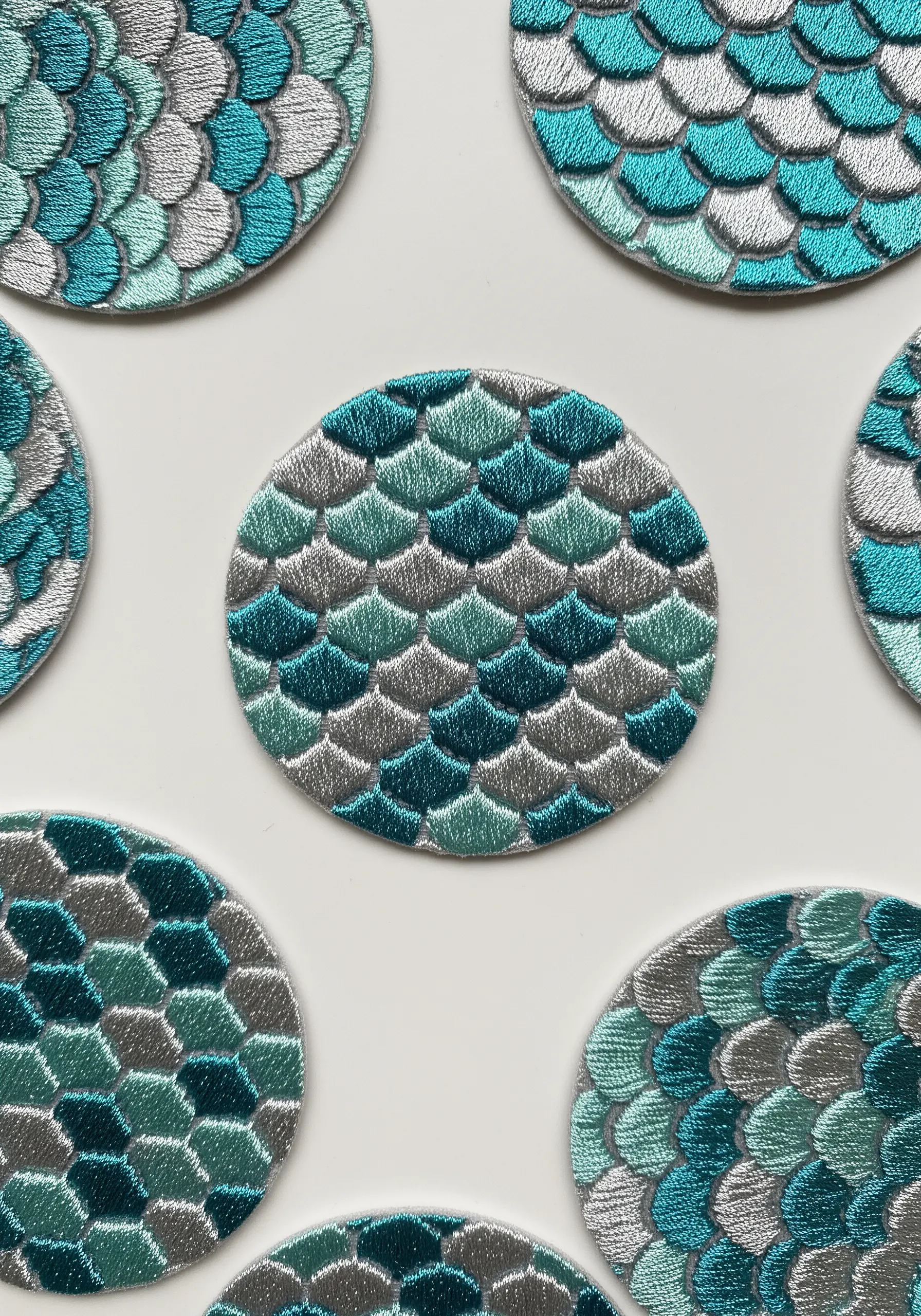 Circular embroidered coasters with a fish scale pattern in variegated teal and silver.