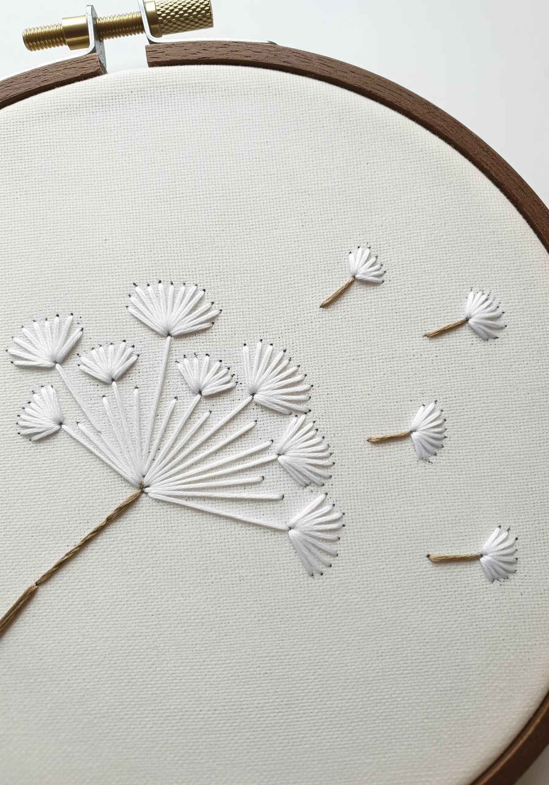 Minimalist embroidered dandelion clock with seeds blowing in the wind on off-white fabric.