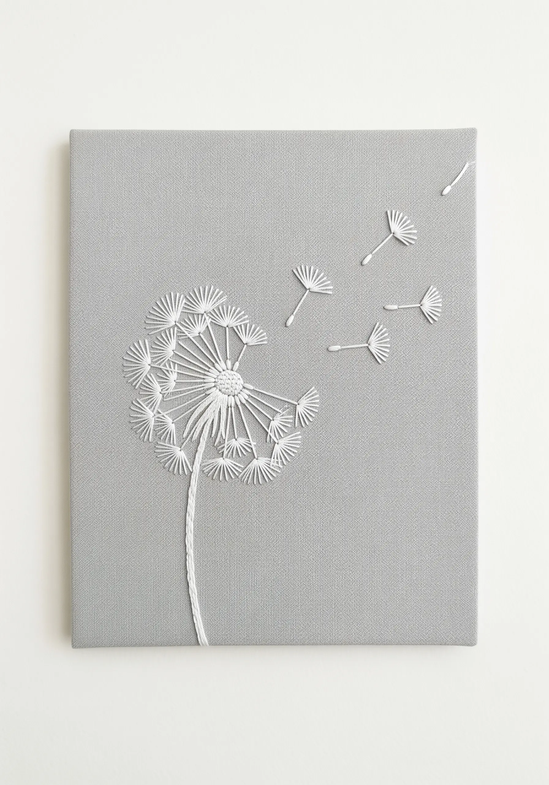 A dandelion with seeds blowing away, embroidered in white on grey canvas.