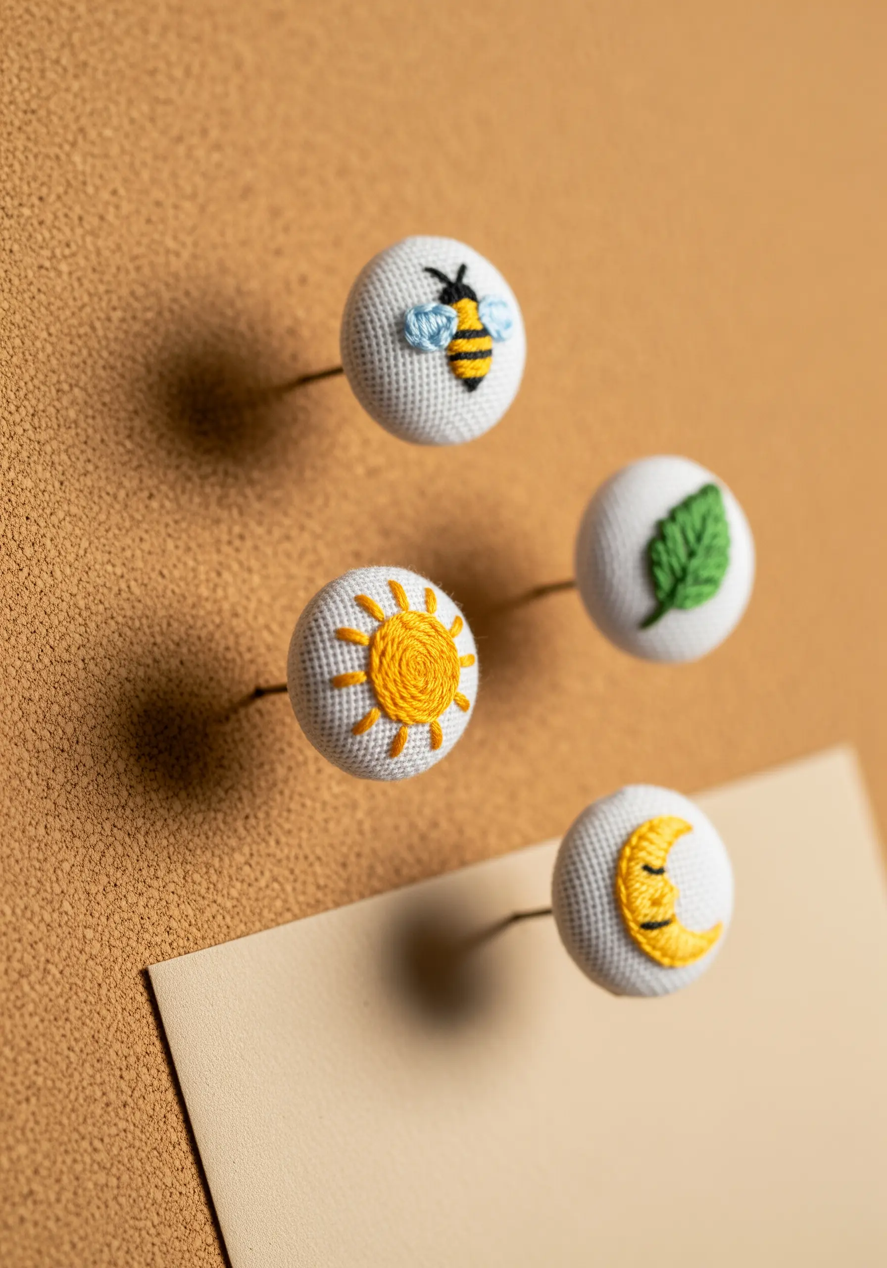 Four handmade push pins with tiny embroidered motifs: a bee, a sun, a leaf, and a moon.