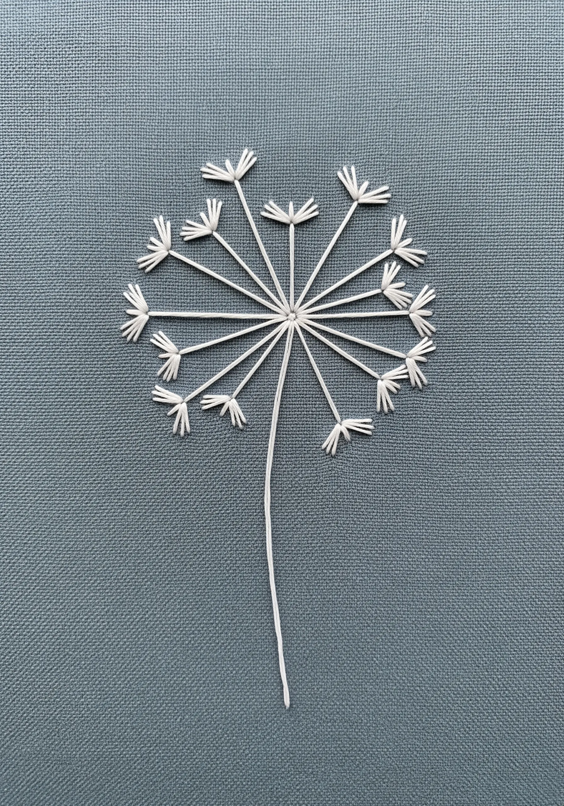 A stylized dandelion seed head embroidered in white thread on a gray-blue fabric.