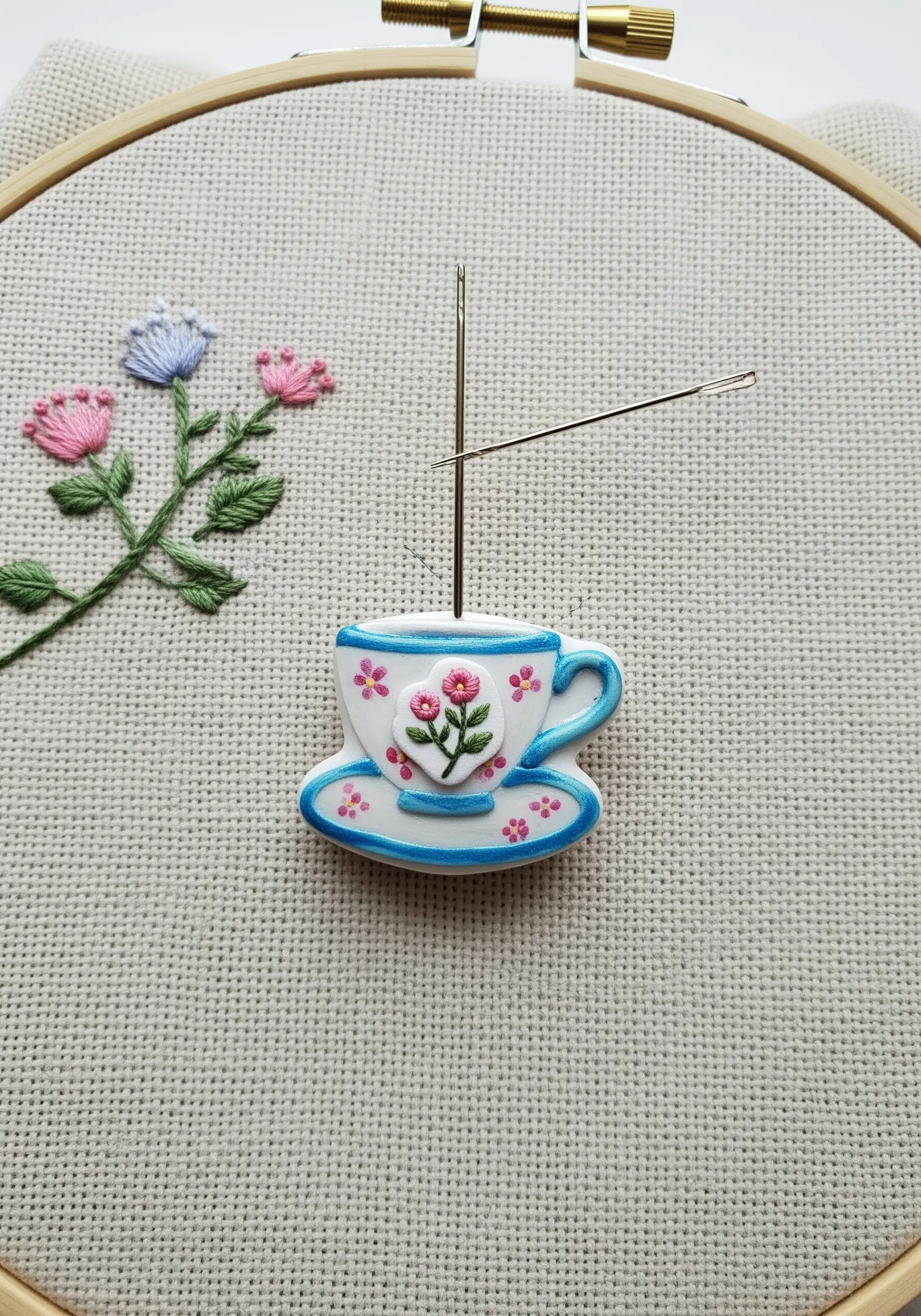 A tiny teacup-shaped needle minder holding two needles on a piece of embroidery fabric