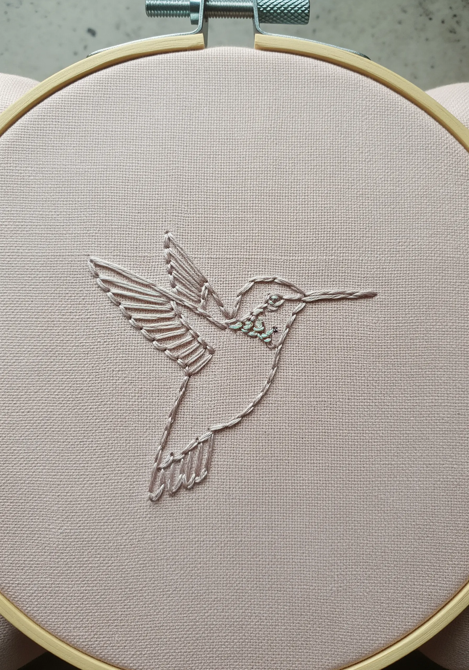 A delicate line-art hummingbird embroidered in white thread on pale pink fabric.