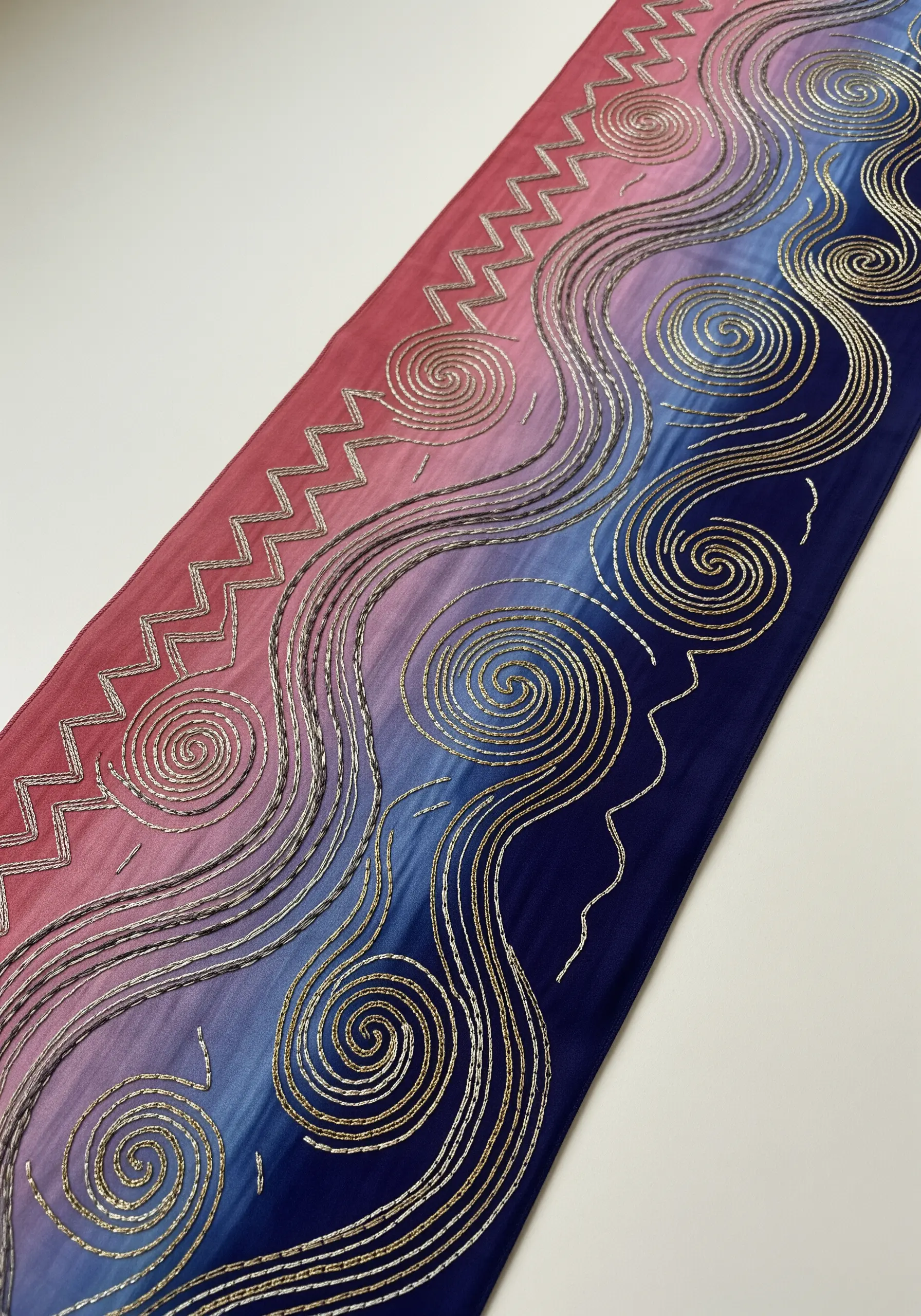 Gold bead embroidery outlining spirals and zigzags on a sheer, pink-to-blue gradient fabric.