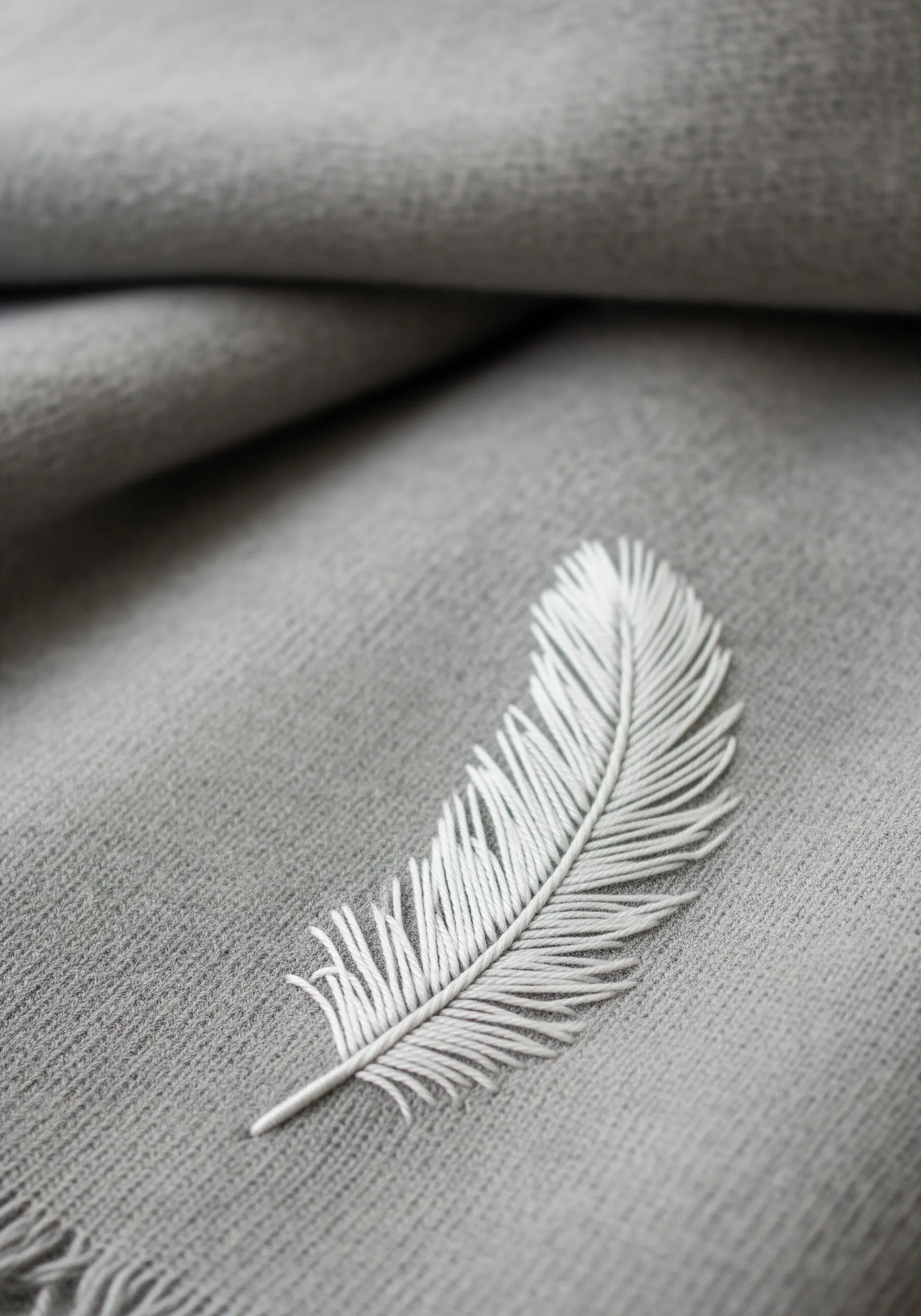 A detailed white embroidered feather with delicate, angled stitches on a grey scarf.