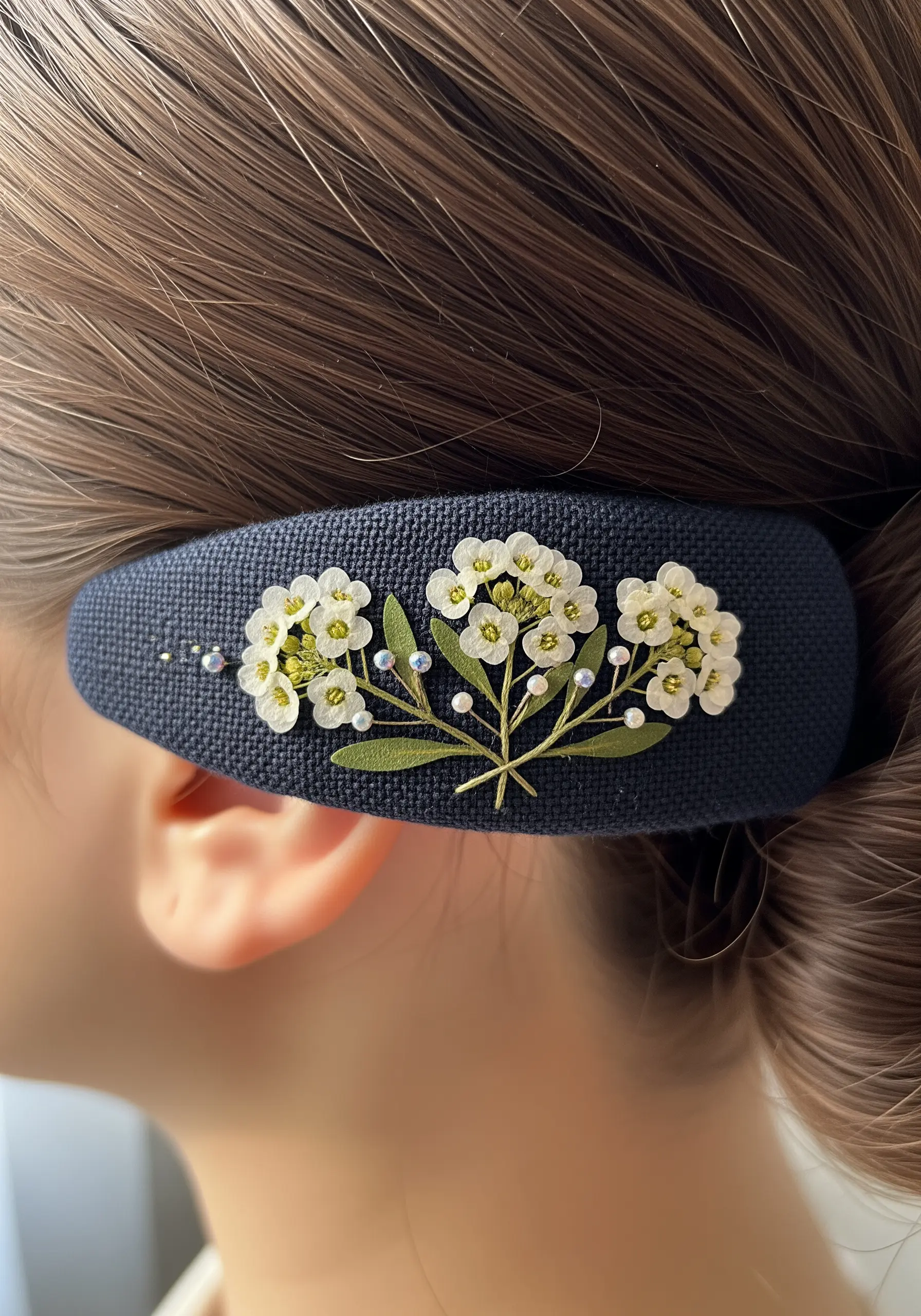 A dark blue hair clip embroidered with tiny pressed white flowers, green leaves, and seed beads.