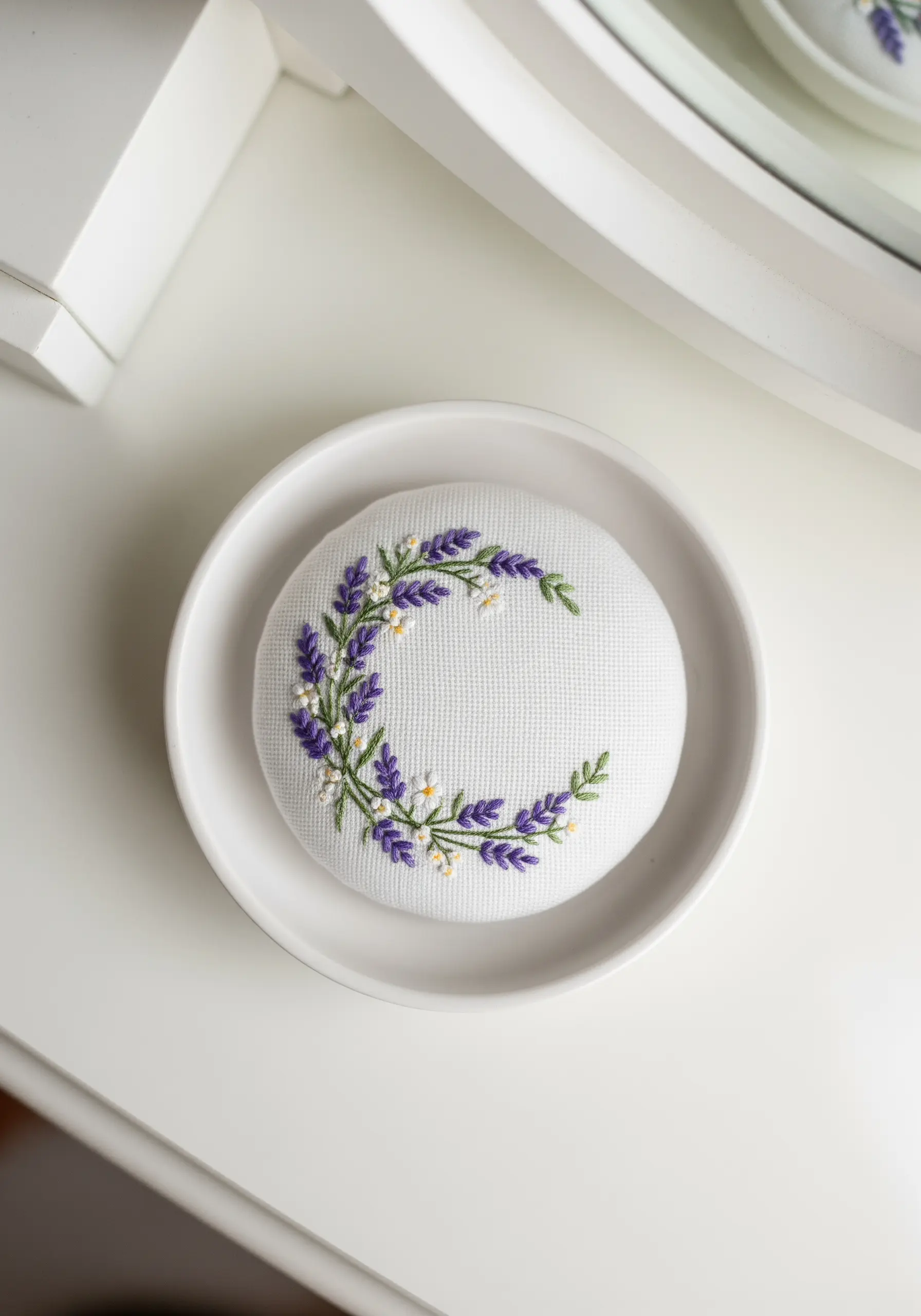 A crescent-shaped wreath of lavender and daisies embroidered on a white pincushion.