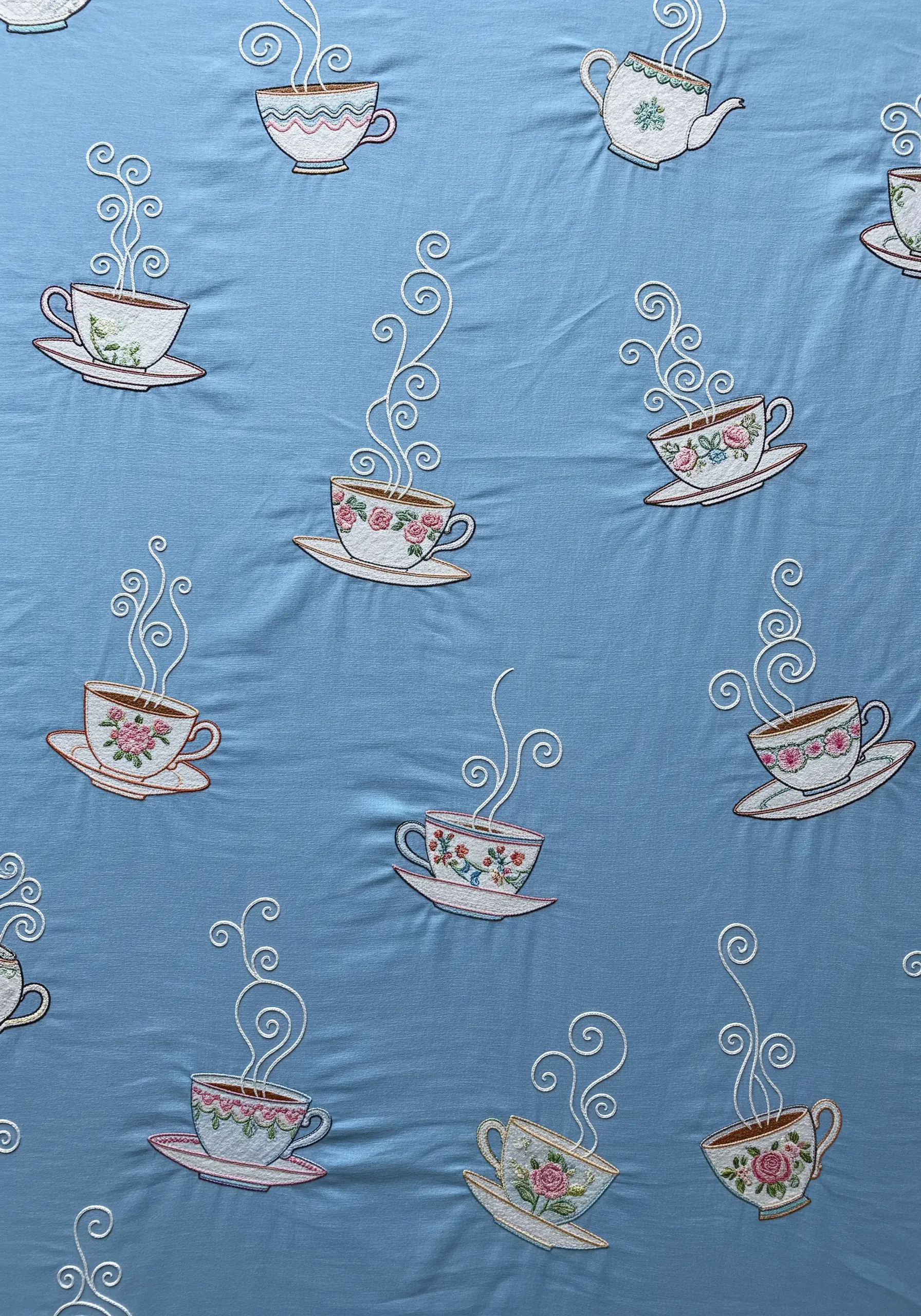 A scatter pattern of various embroidered teacups with swirling steam on blue fabric.