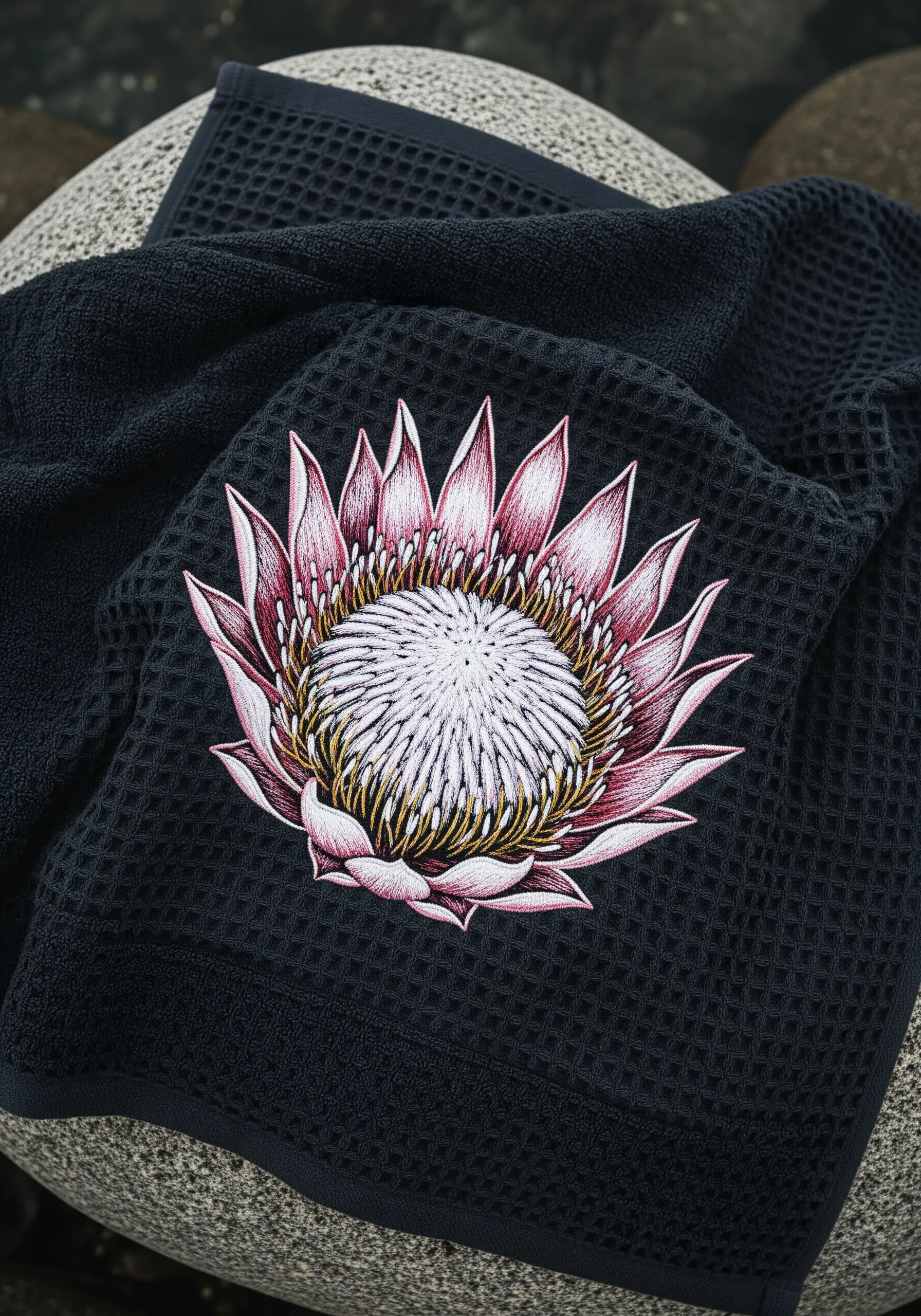 A detailed pink and white King Protea flower embroidered on a dark grey waffle-weave towel.