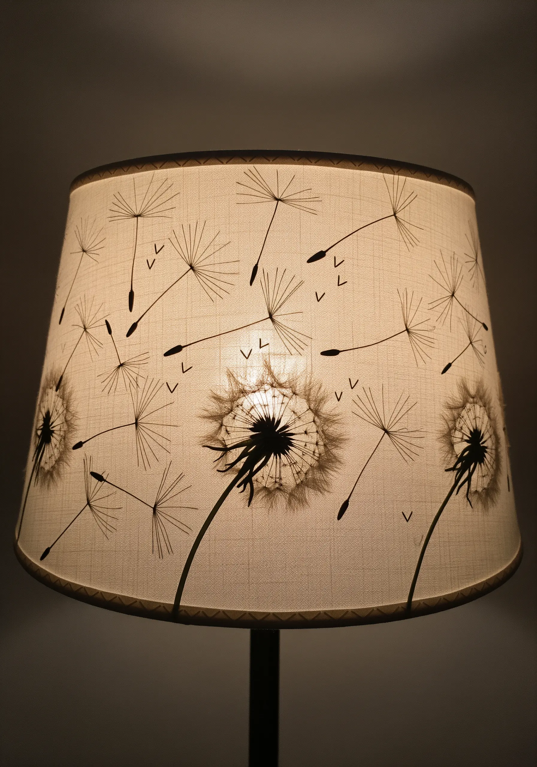 A fabric lampshade illuminated from within, showing an embroidered pattern of dandelion seeds.