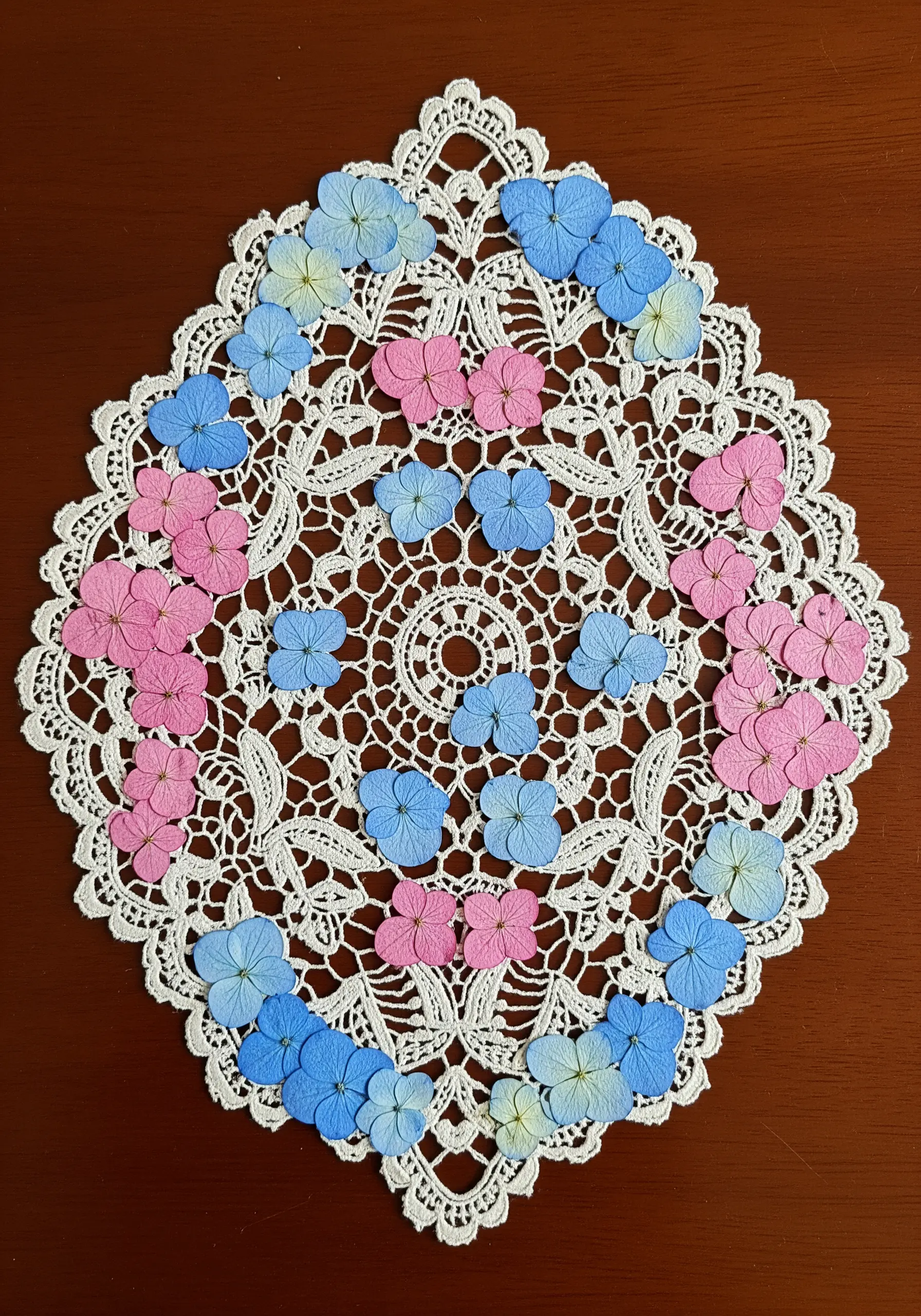 A white lace doily decorated with small, colorful pressed hydrangea petals arranged in a pattern.