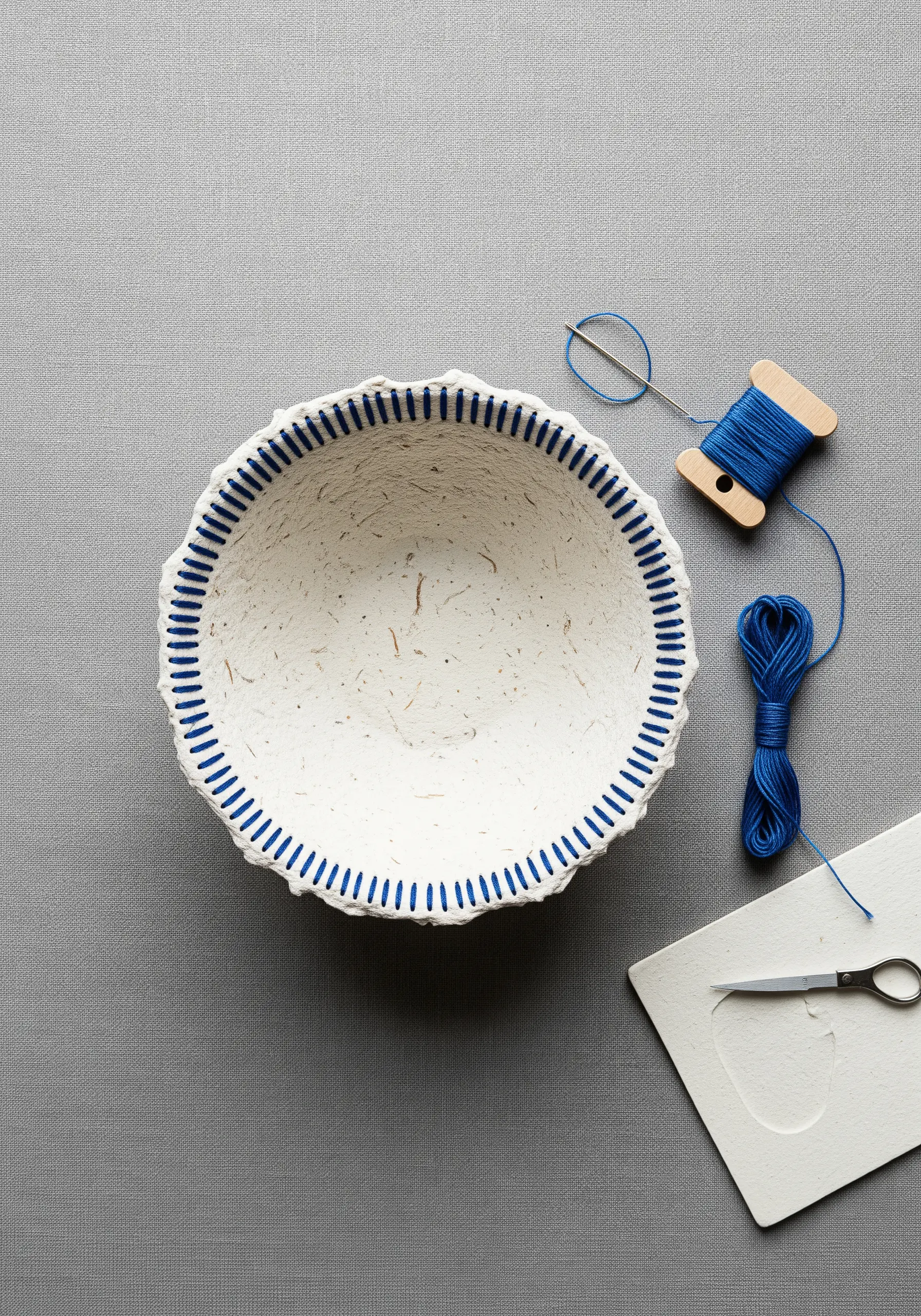 A white papier-mâché bowl with a border of blue vertical stitches around the rim.