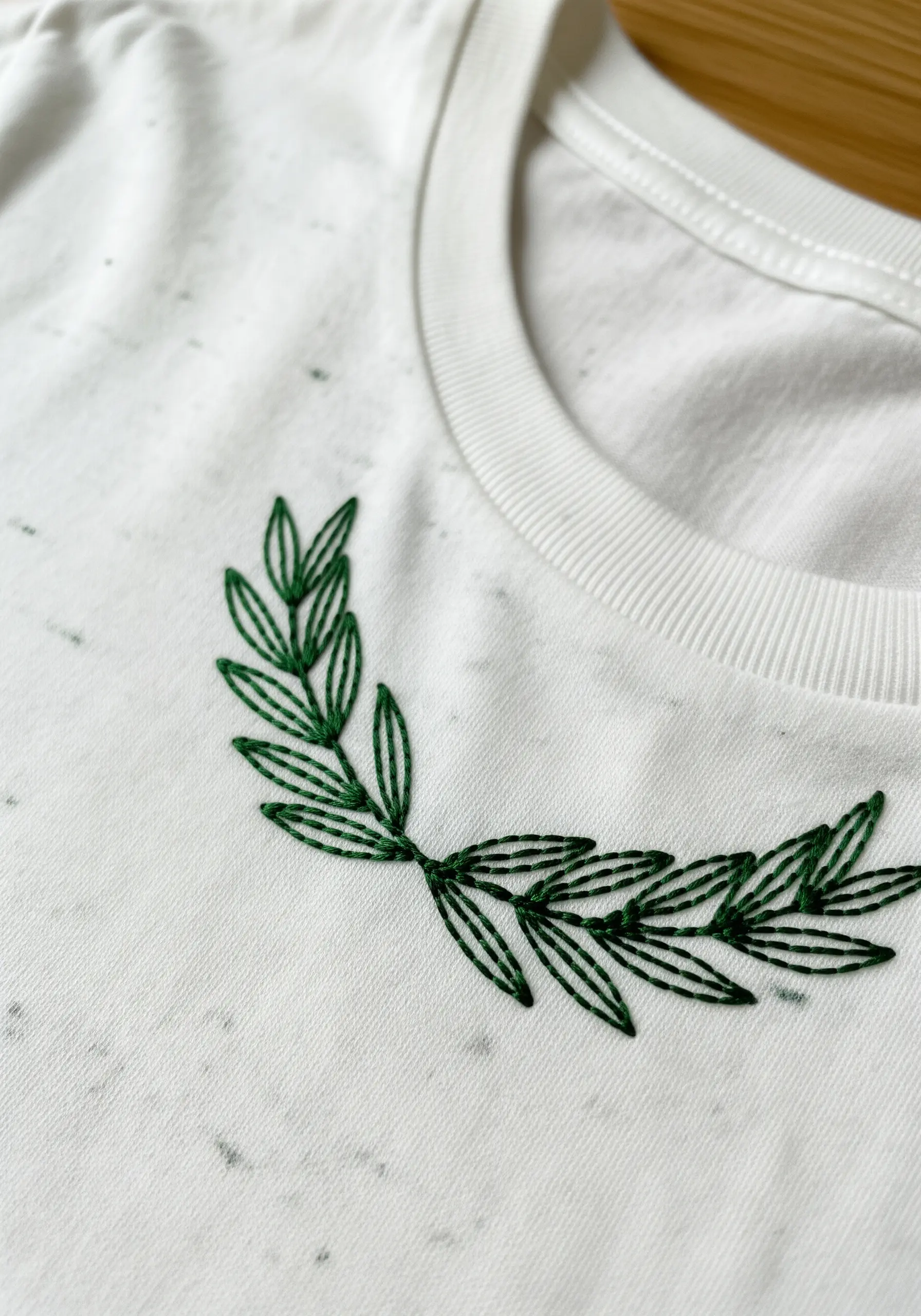 Green laurel branch outline stitch embroidery on a white speckled t-shirt collar.