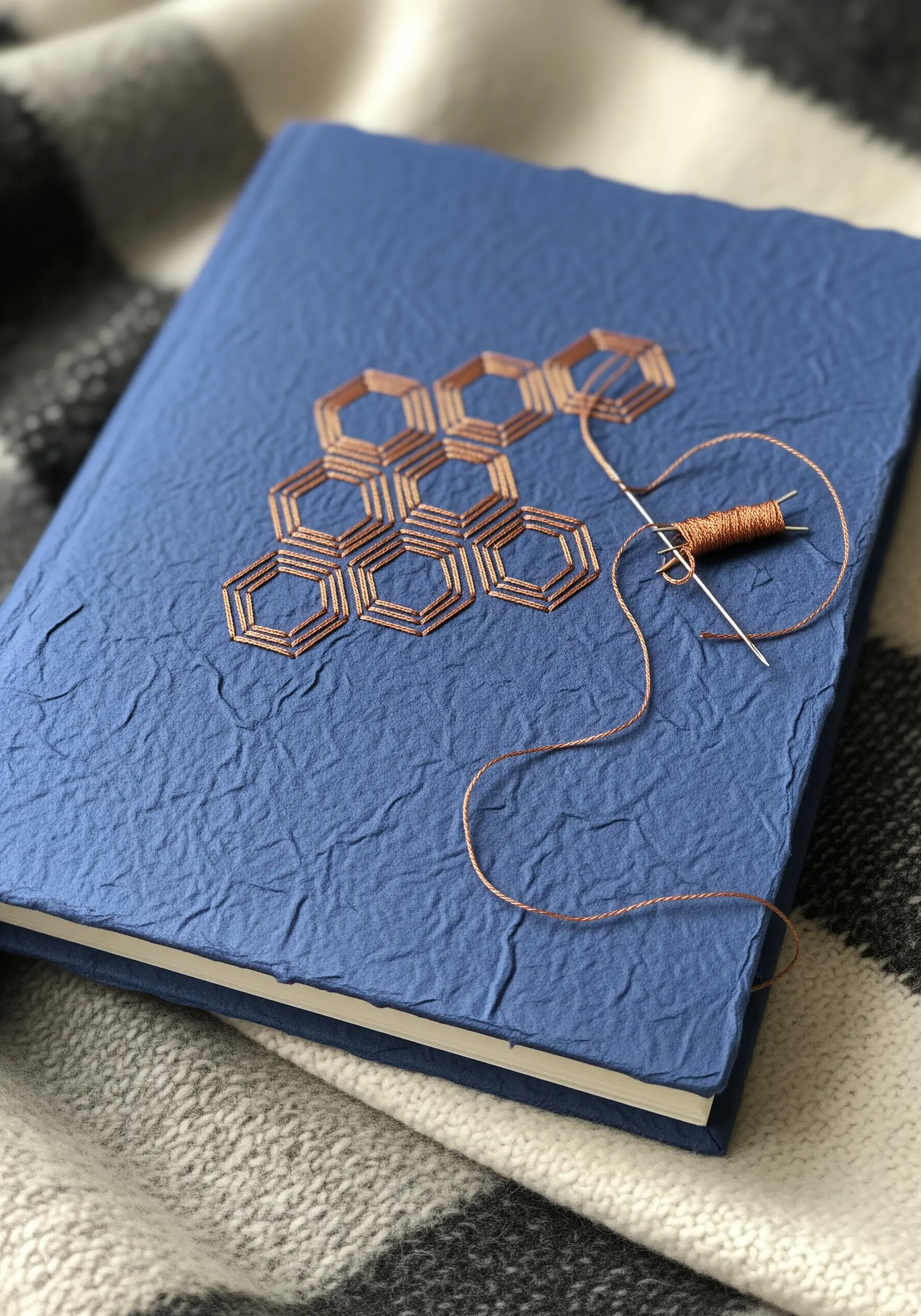 A blue textured paper journal cover being embroidered with a copper-colored geometric honeycomb pattern.