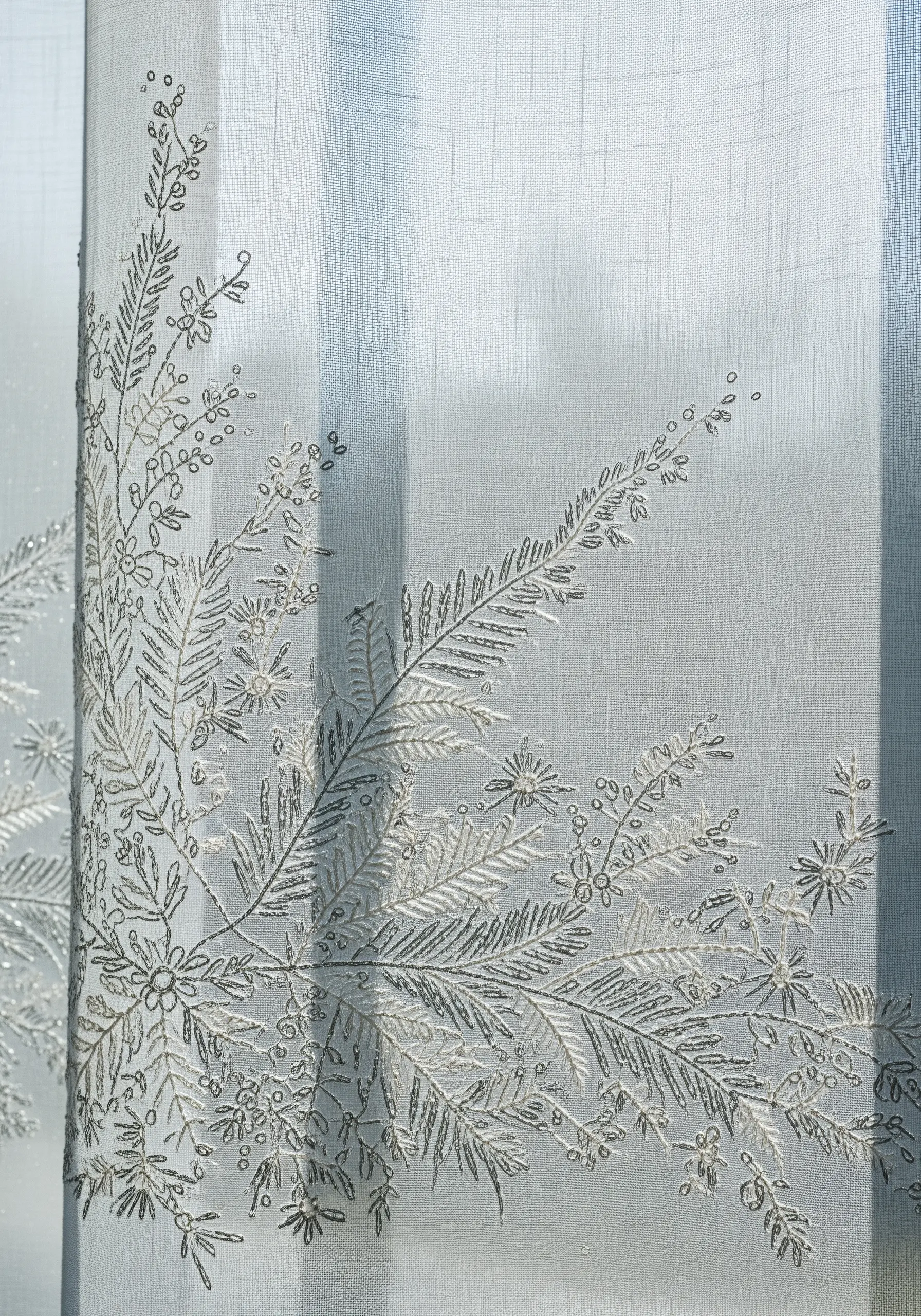 Delicate white and silver embroidery of frosty branches on a sheer, light blue curtain.