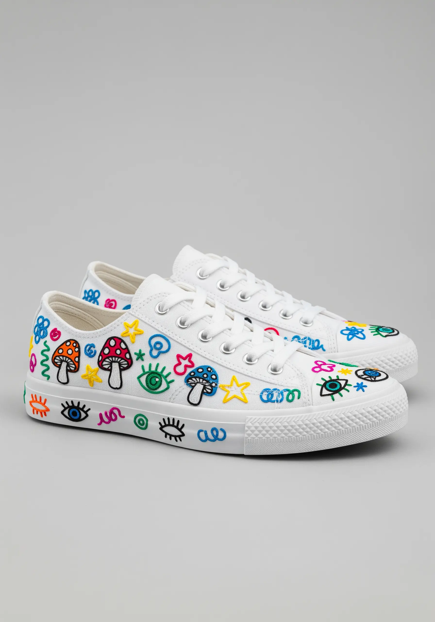 White canvas sneakers covered in colorful, doodle-style embroidered motifs like mushrooms and eyes.