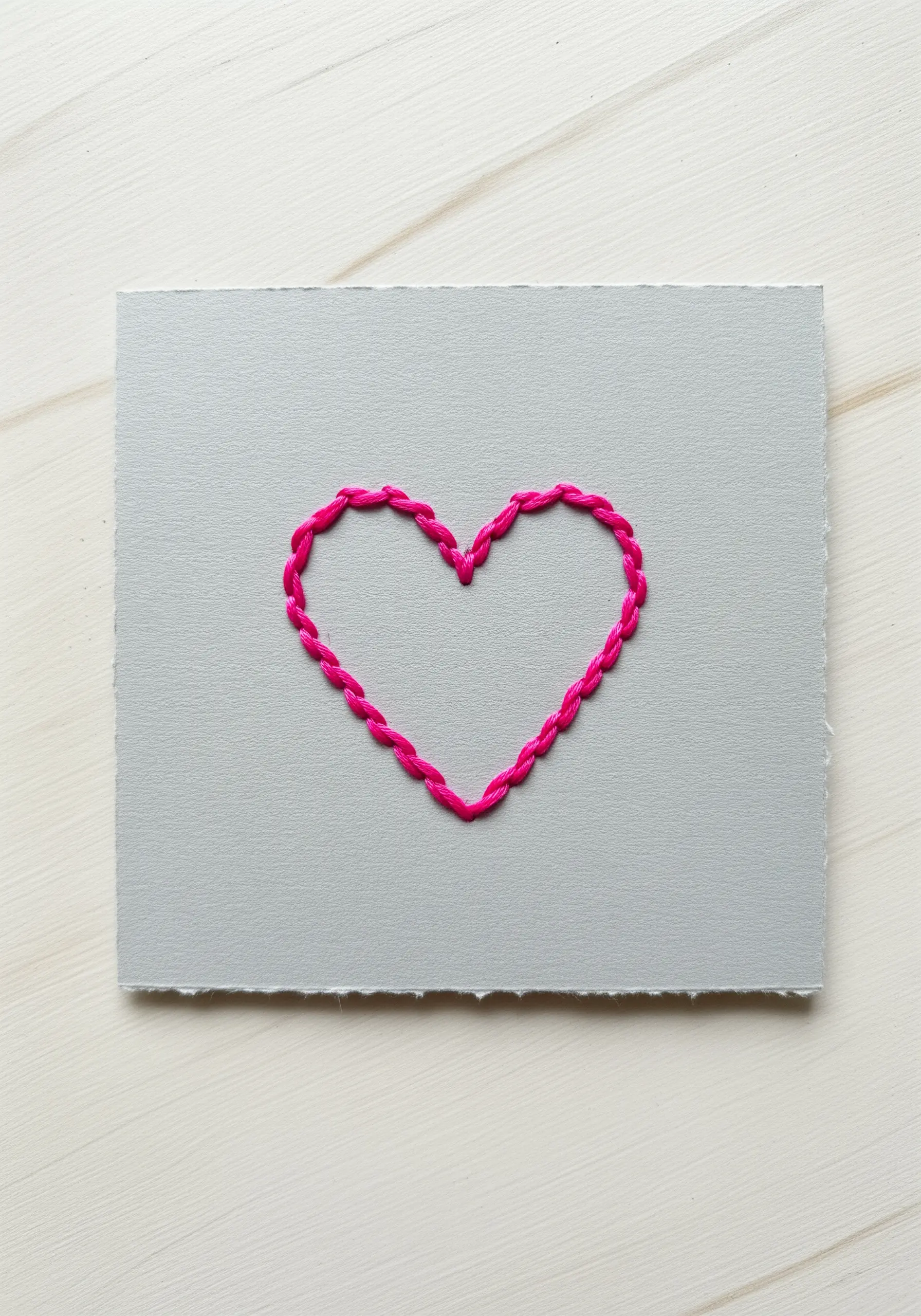A bright pink chain stitch heart embroidered onto a piece of grey textured paper.