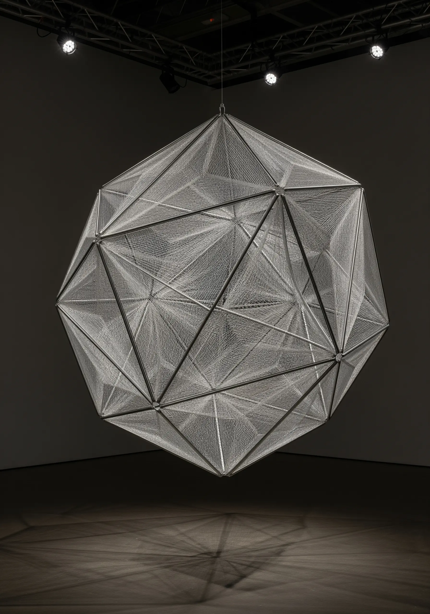 A large, complex metal polyhedron suspended in a dark room, intricately laced with fine white thread.