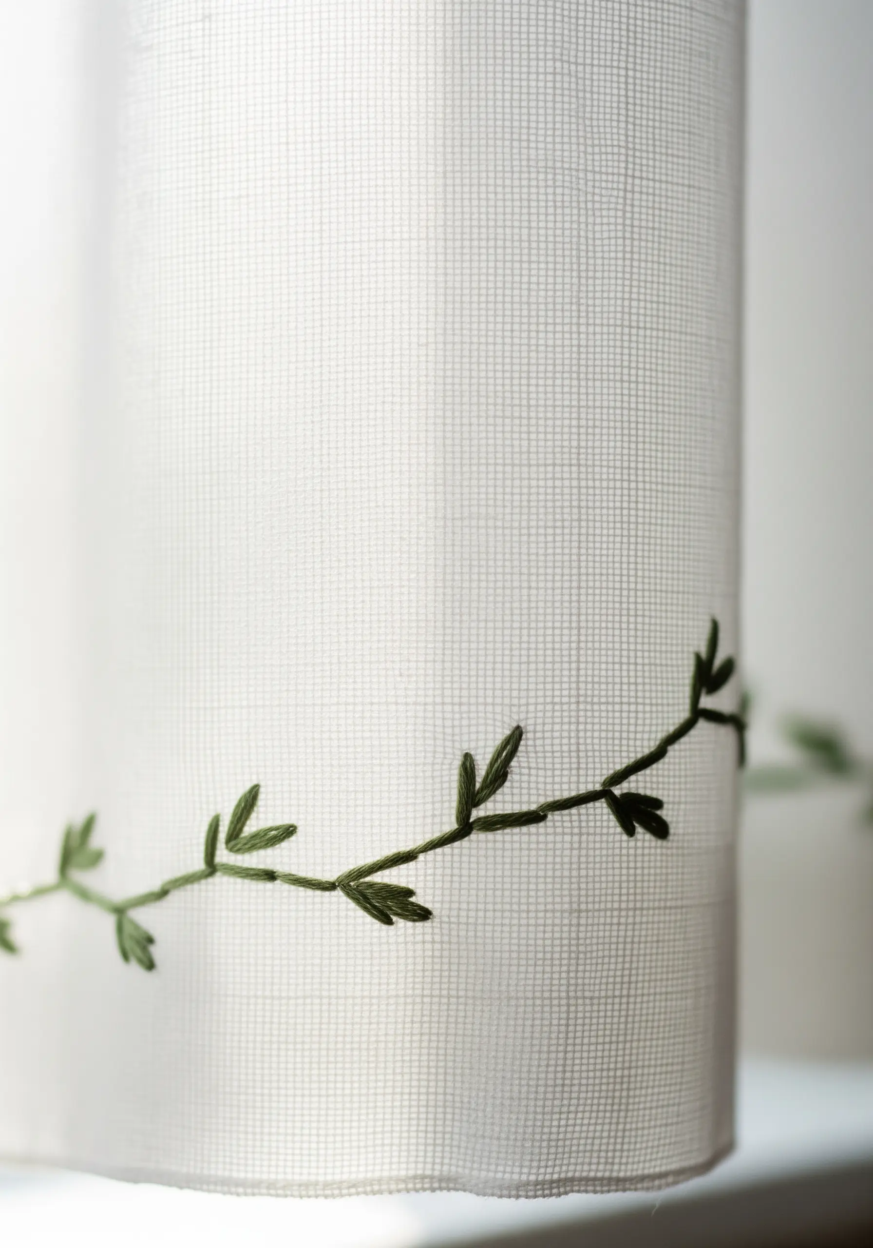 A delicate green vine with leaves embroidered along the bottom of a white sheer curtain.