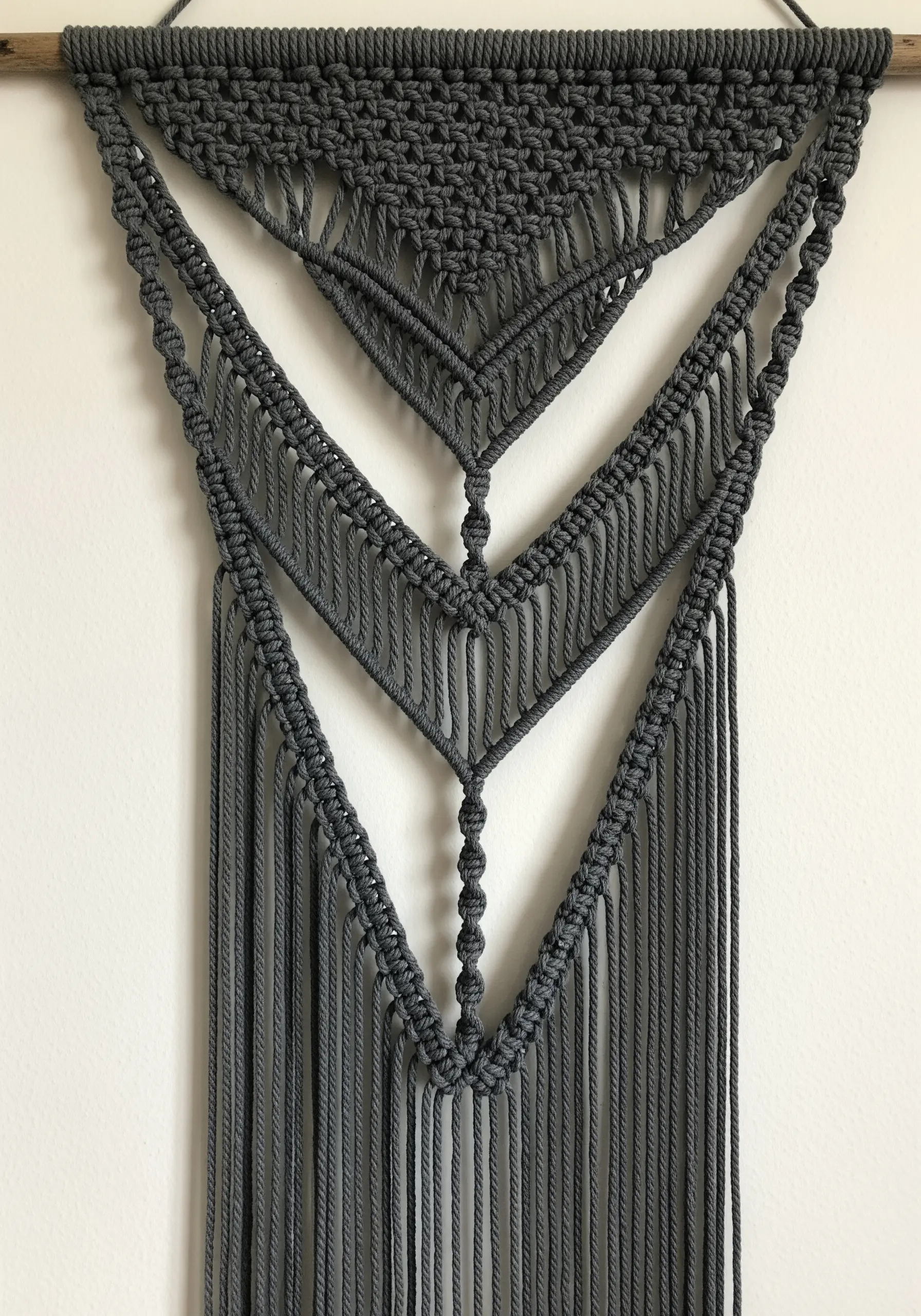 A close-up of a dark gray macrame wall hanging with intricate, layered geometric patterns.