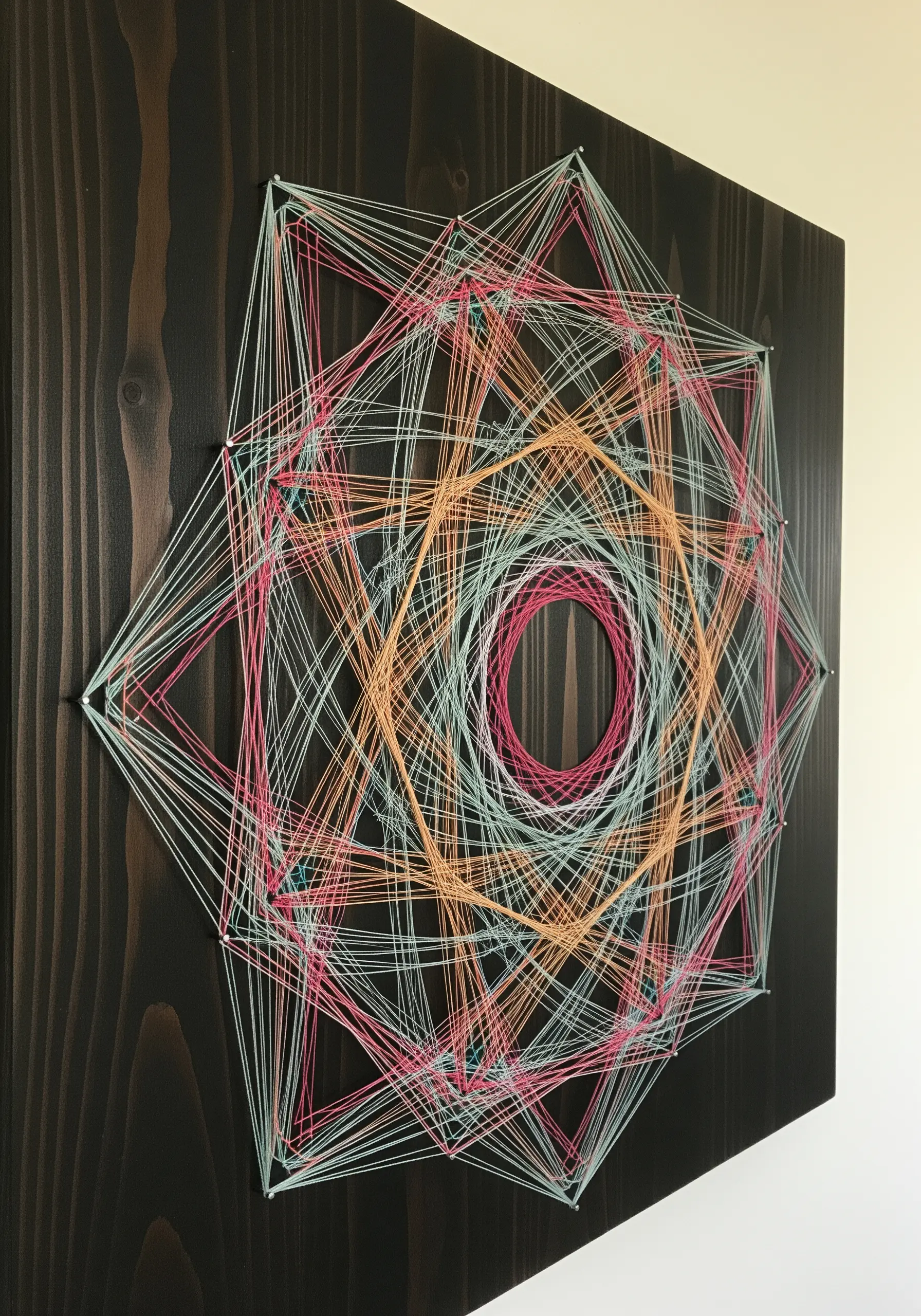 Geometric string art on a dark wood panel, with intricate layers of pink, orange, and blue thread.