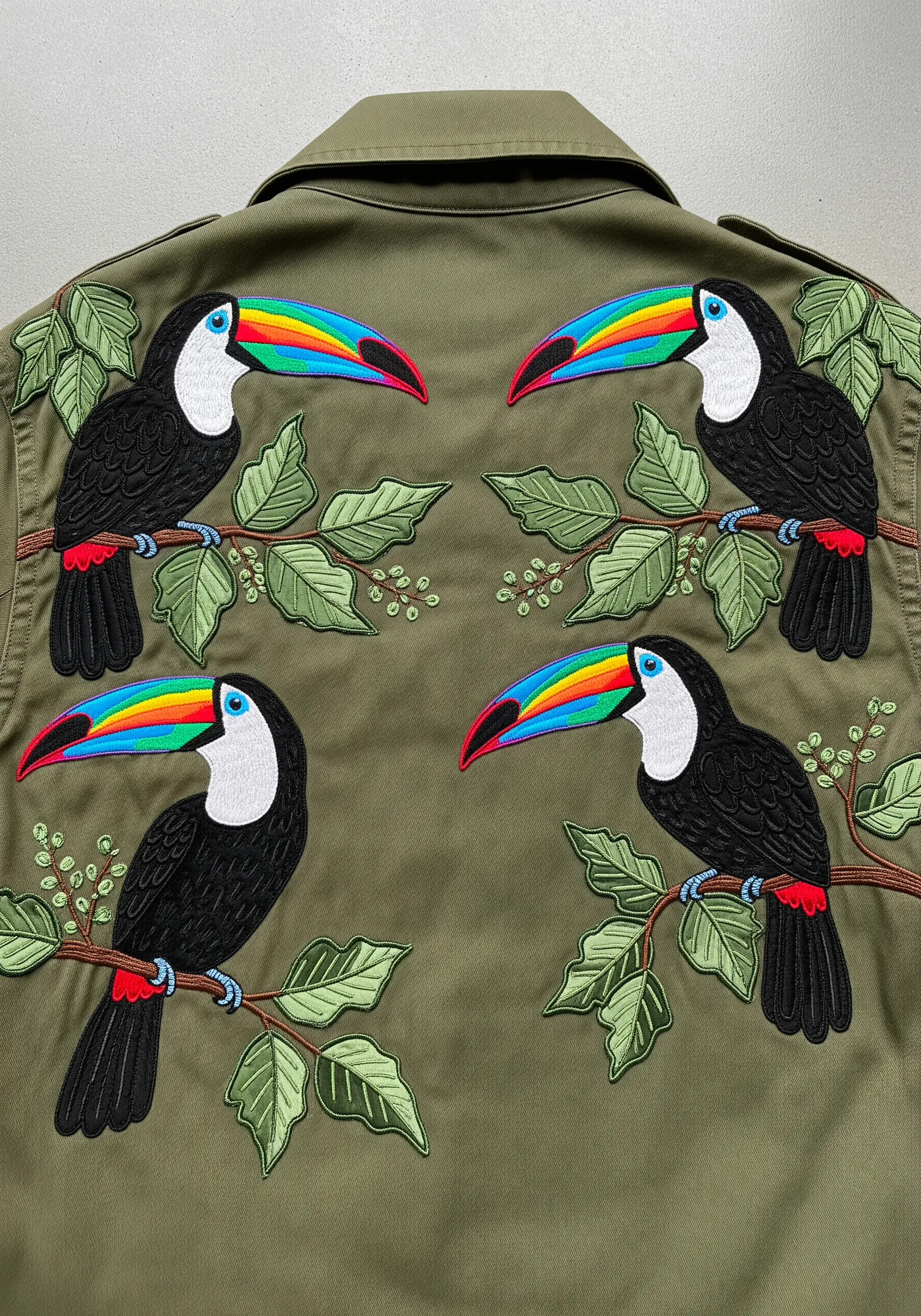 Four toucans arranged in a symmetrical, mirrored pattern on the back of an olive green jacket.
