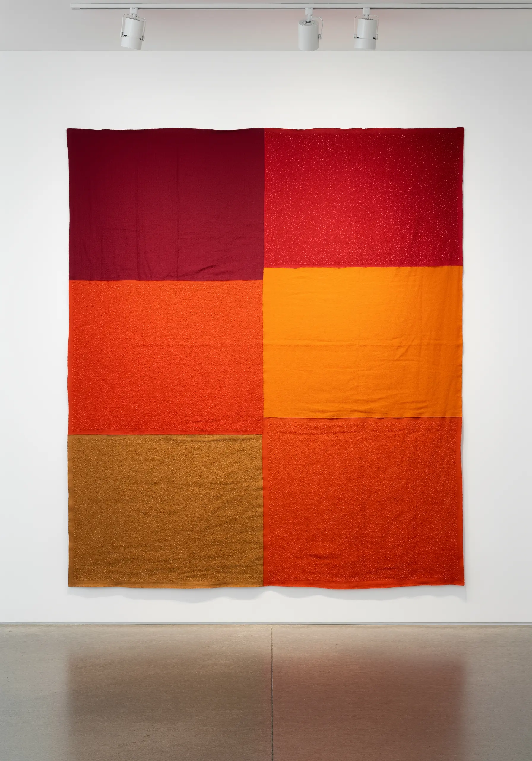 Large textile wall art with blocks of red, orange, and brown fabric with subtle textural stitching.