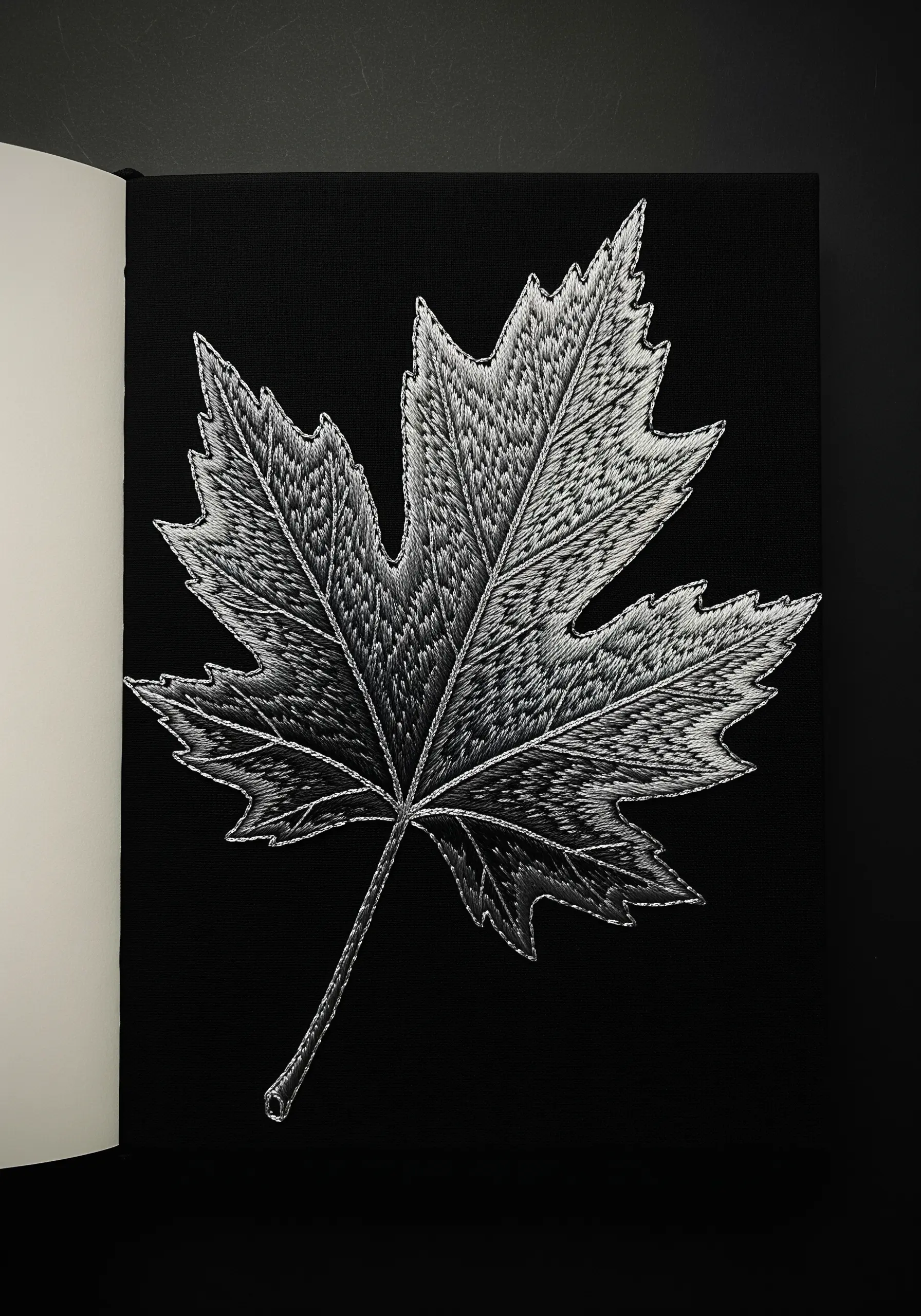 A highly detailed, thread-painted maple leaf in black, white, and grey on a black journal.