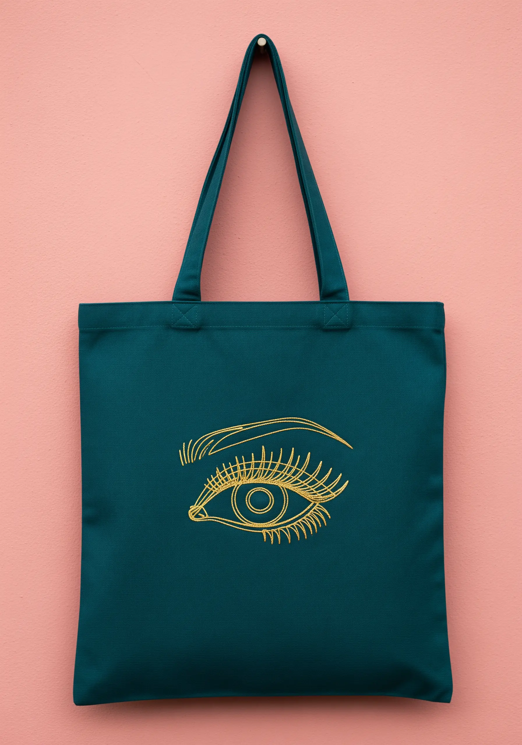 A detailed line art eye embroidered in gold thread on a teal tote bag.