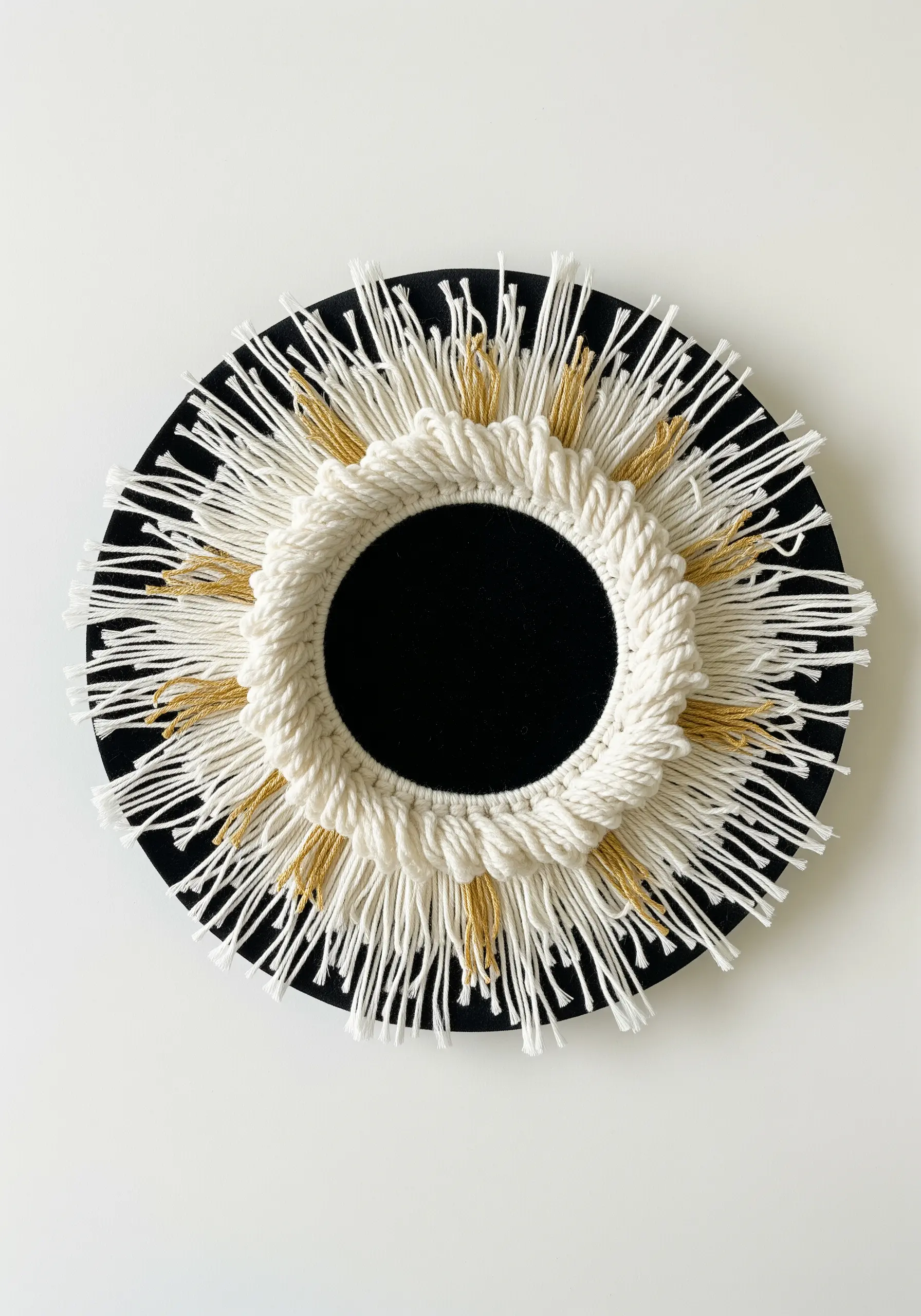 A macrame sunburst design around a black circular center, with white and gold threads.