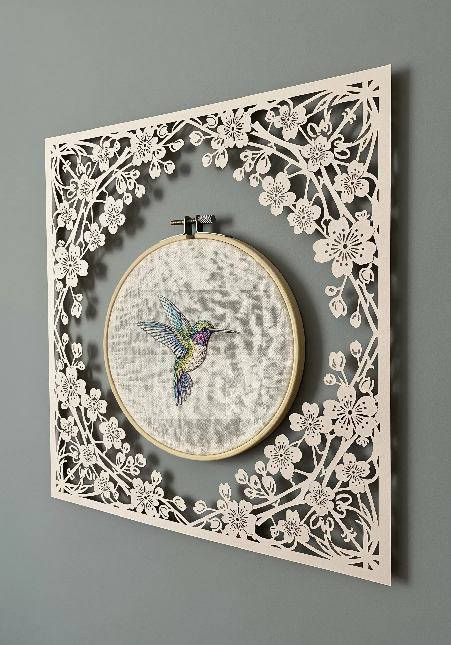 Embroidered hummingbird in a hoop, framed by a square white paper-cut floral design.