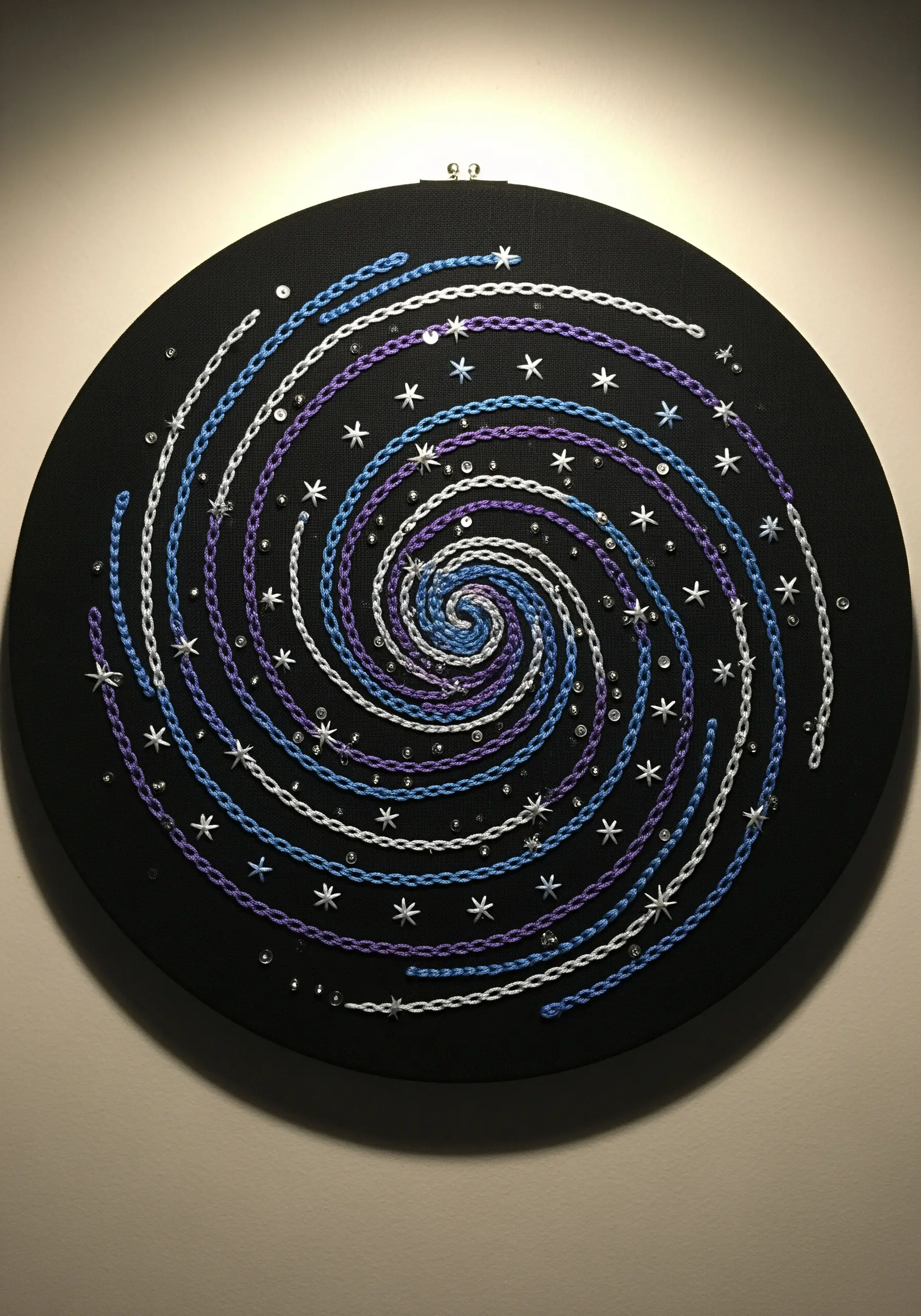 A spiral galaxy mandala made of chain stitches in blue, purple, and white, with scattered sequins.