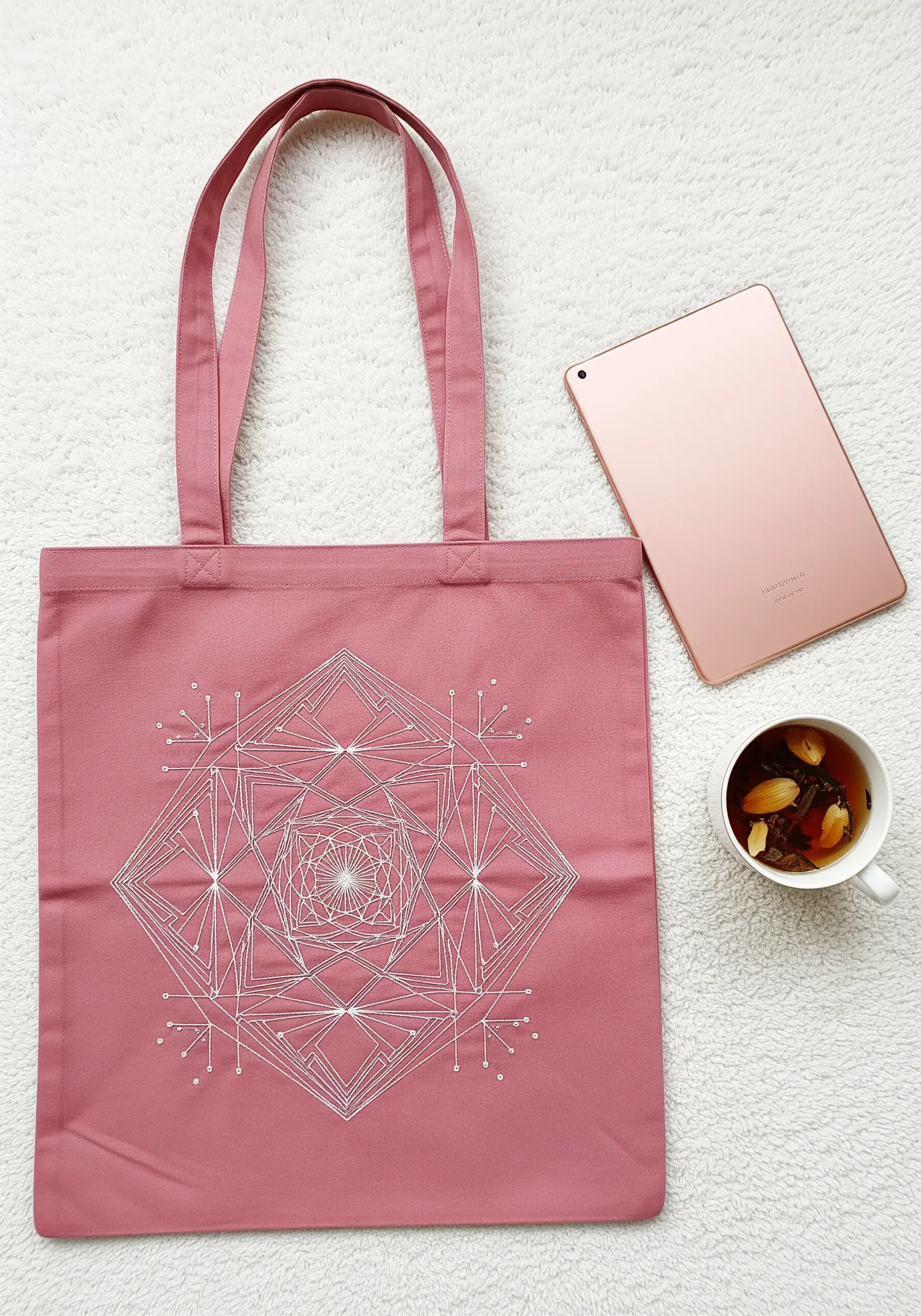 A complex geometric mandala embroidered in white thread on a dusty rose tote.
