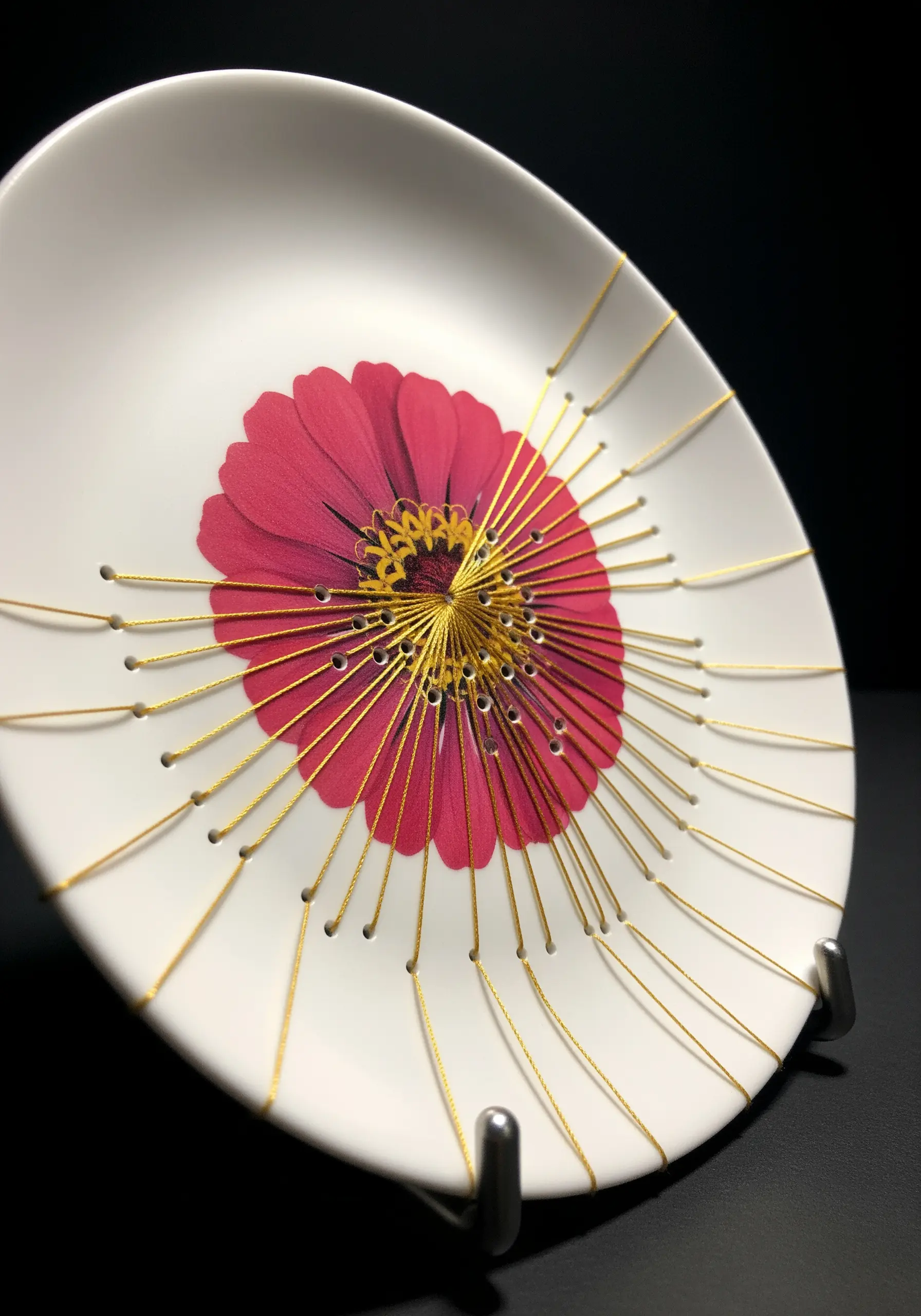 A pressed pink flower on a white plate, surrounded by radiating gold string art.