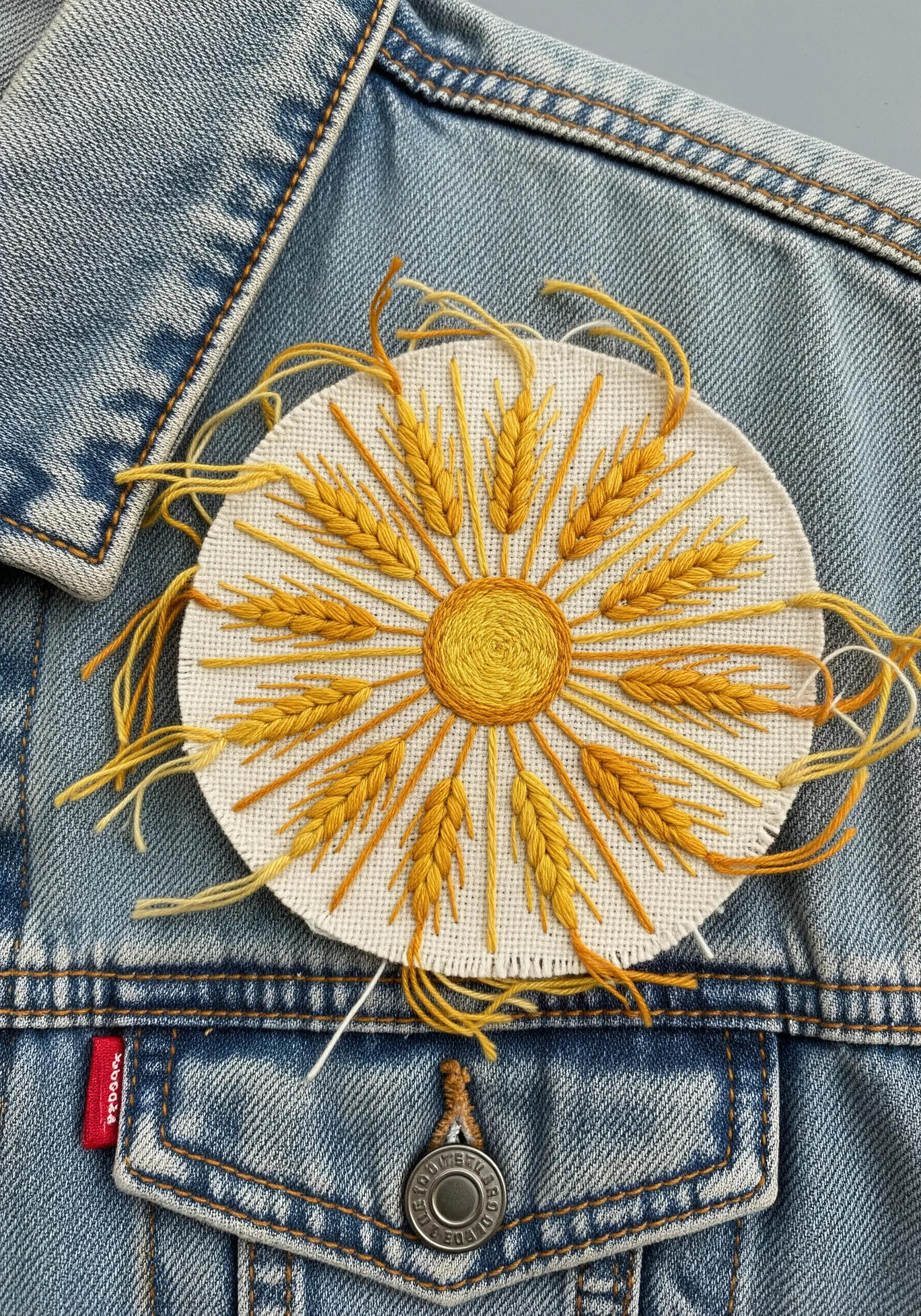 An embroidered patch of a sun made from wheat stalks, ready to be sewn onto a denim jacket.