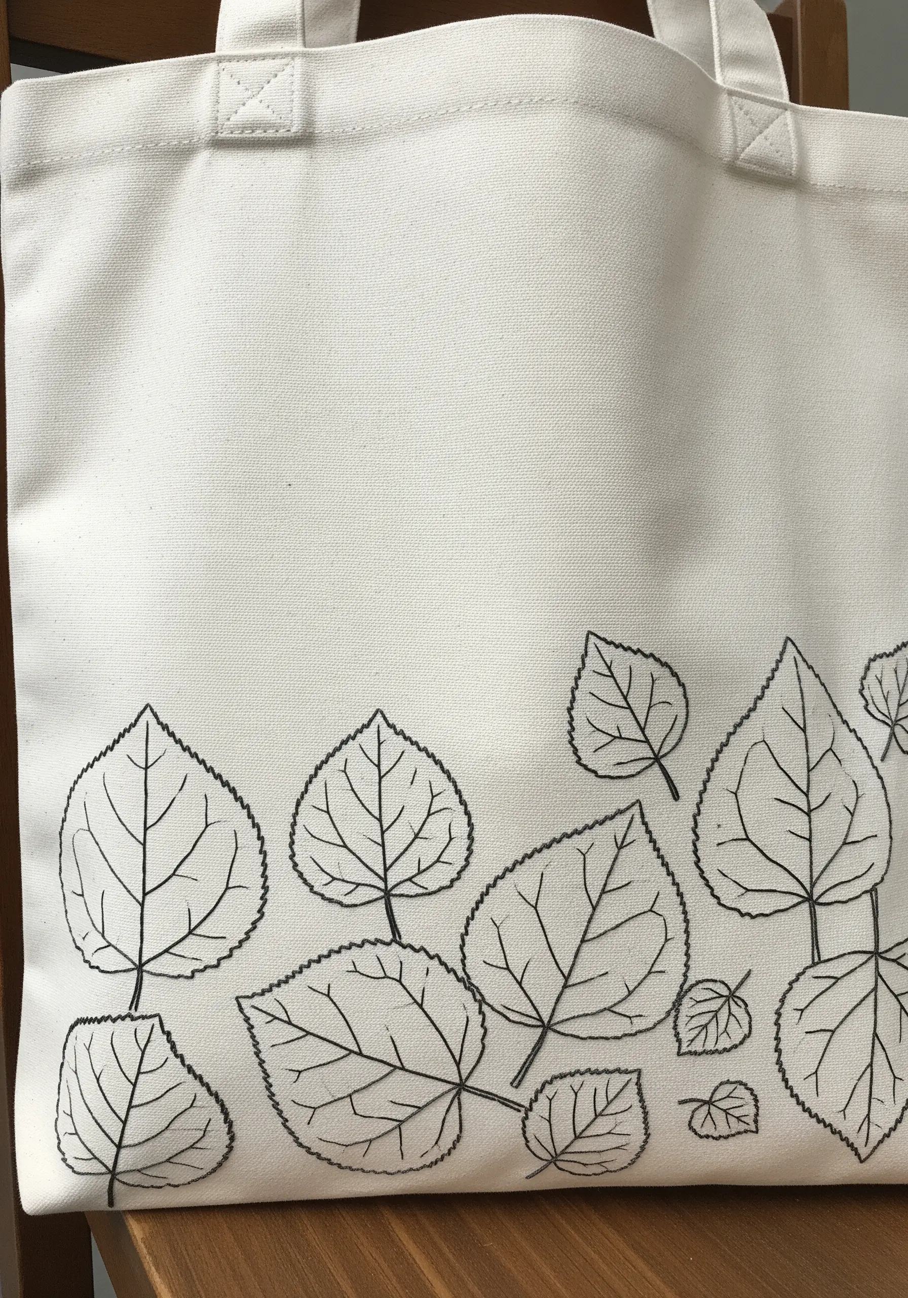A canvas tote bag decorated with a border of clean, black backstitch leaf outlines.