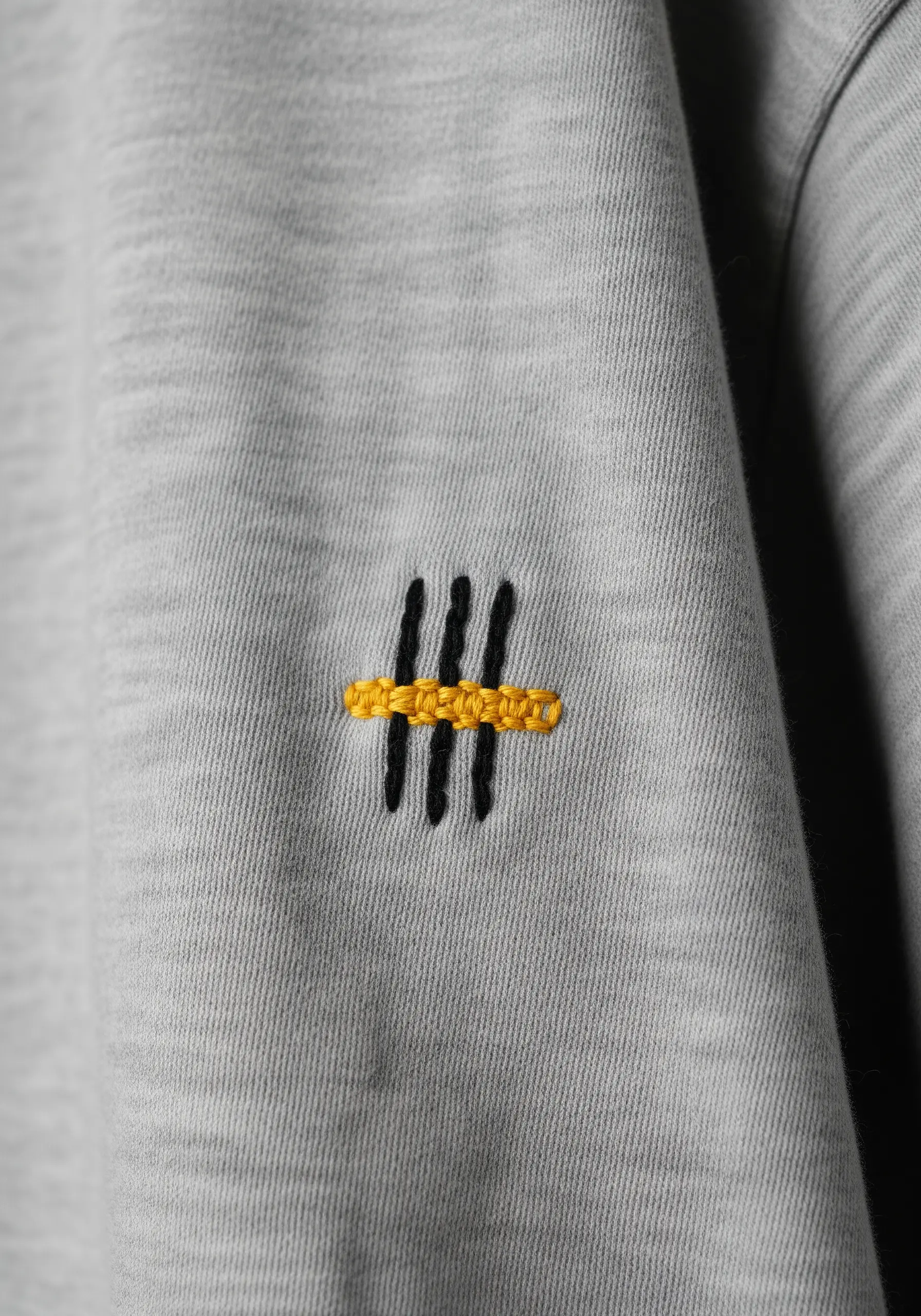 Three vertical black stitches crossed by a textured yellow horizontal bar on a grey sleeve.