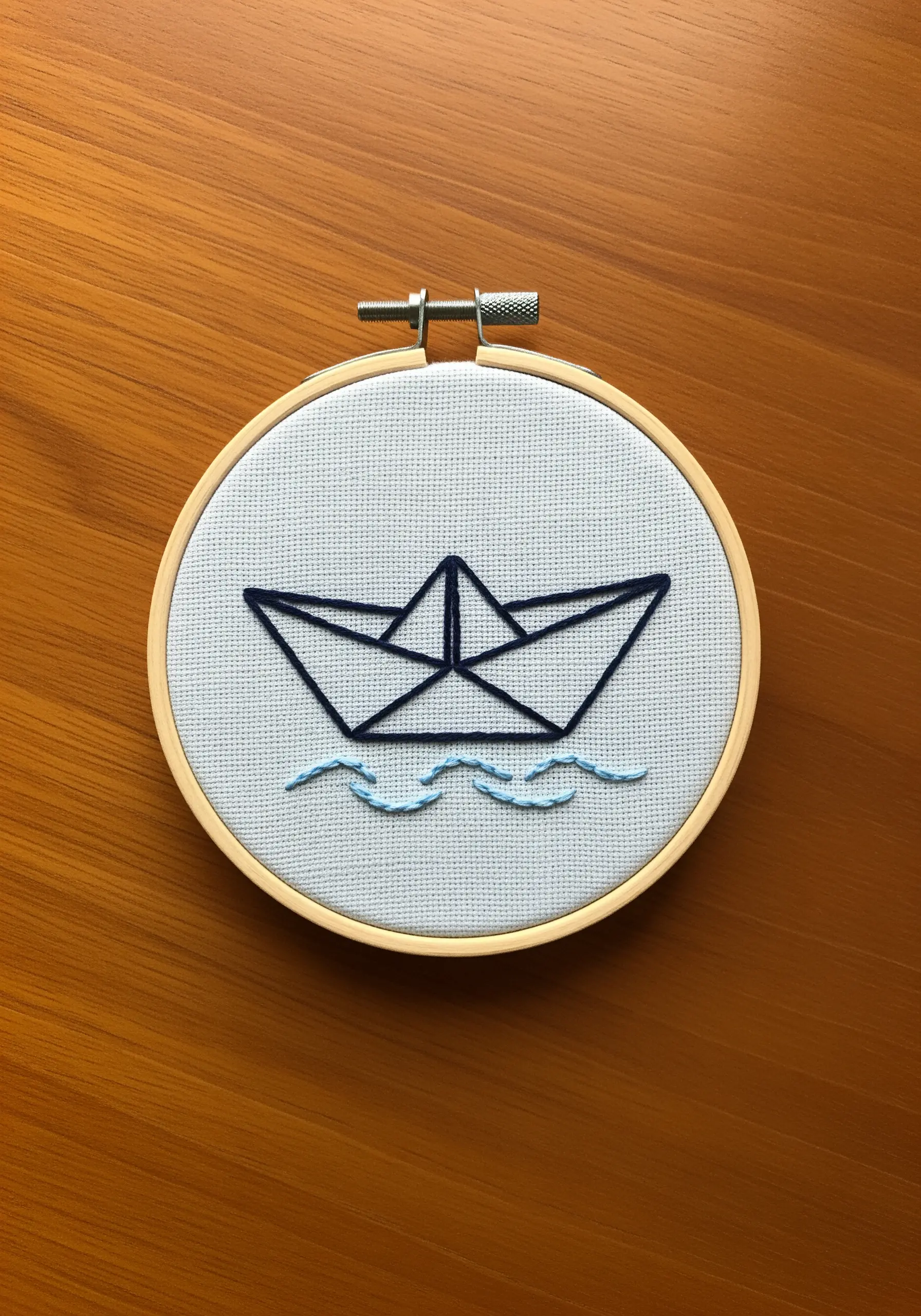 A minimalist paper boat outlined in dark blue thread, with light blue wavy lines below.