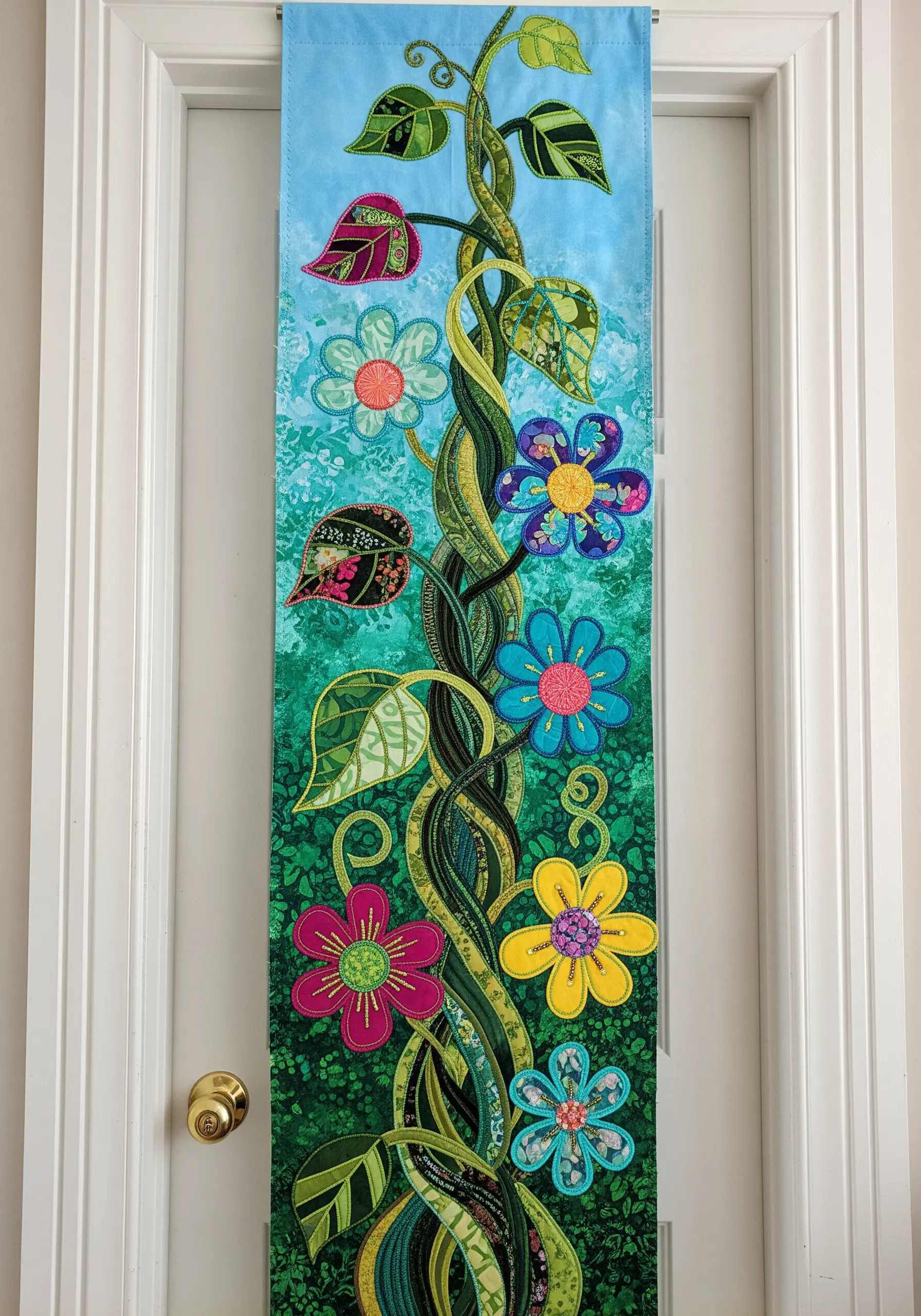 A tall wall hanging with a climbing vine and colorful appliqué flowers on a patterned background.