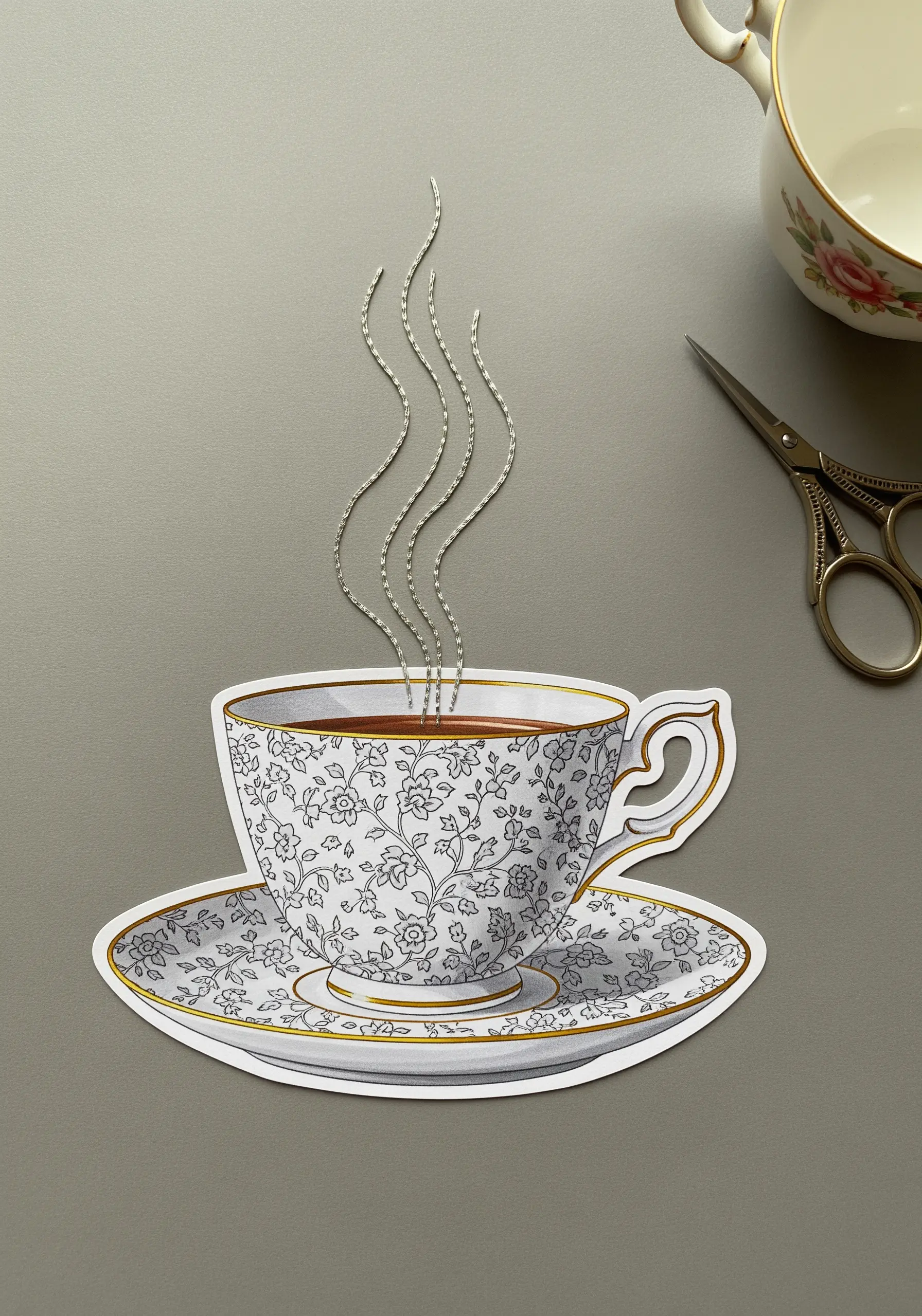 A paper cutout of a floral teacup with wavy lines of silver thread stitched above it as steam.