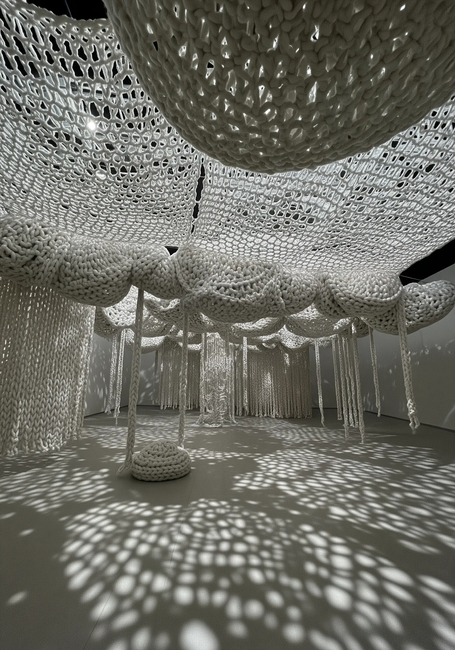 A room installation with a ceiling covered in white, large-scale crocheted clouds and webs.