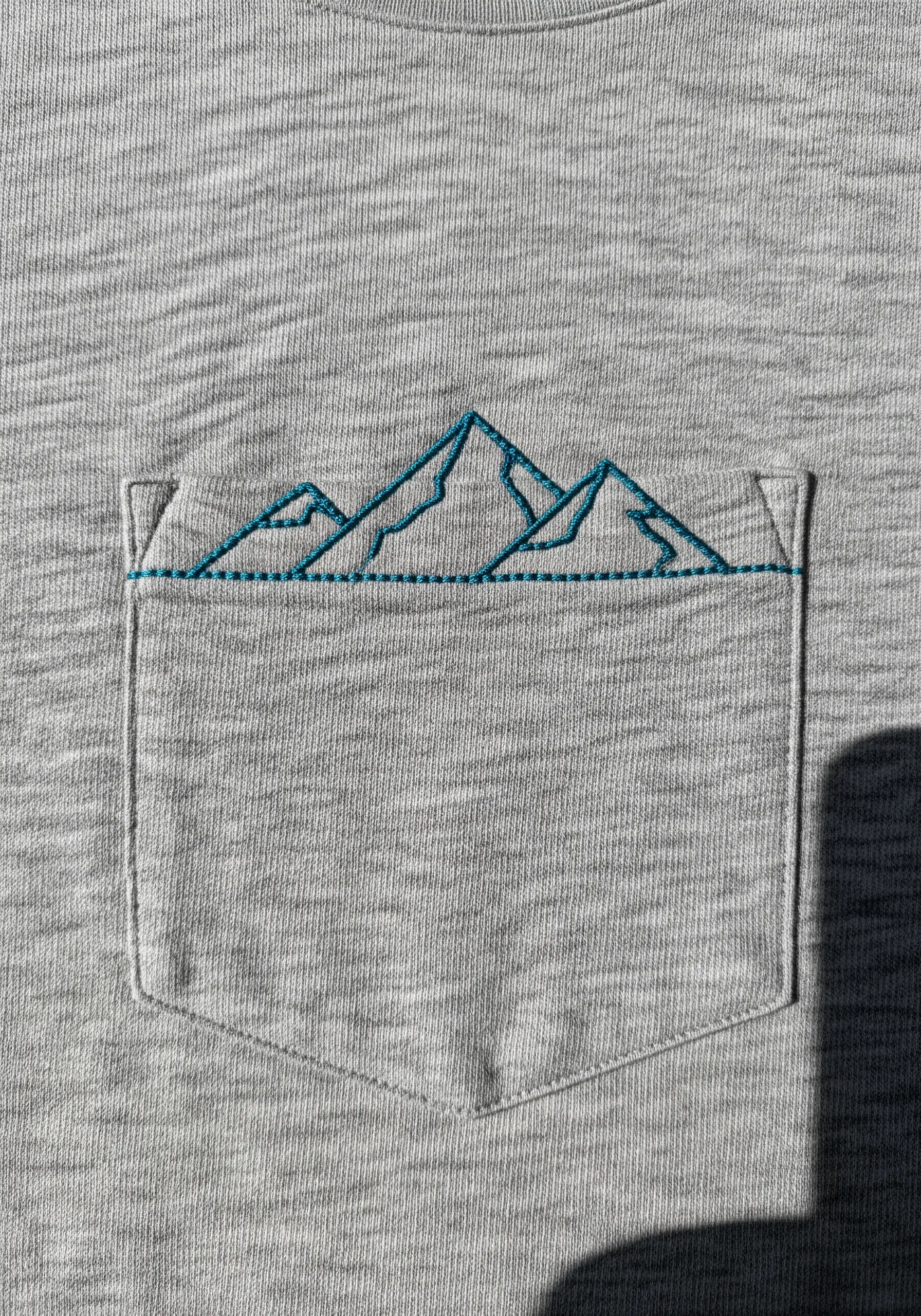 A simple teal outline of a mountain range stitched along the top of a t-shirt pocket.