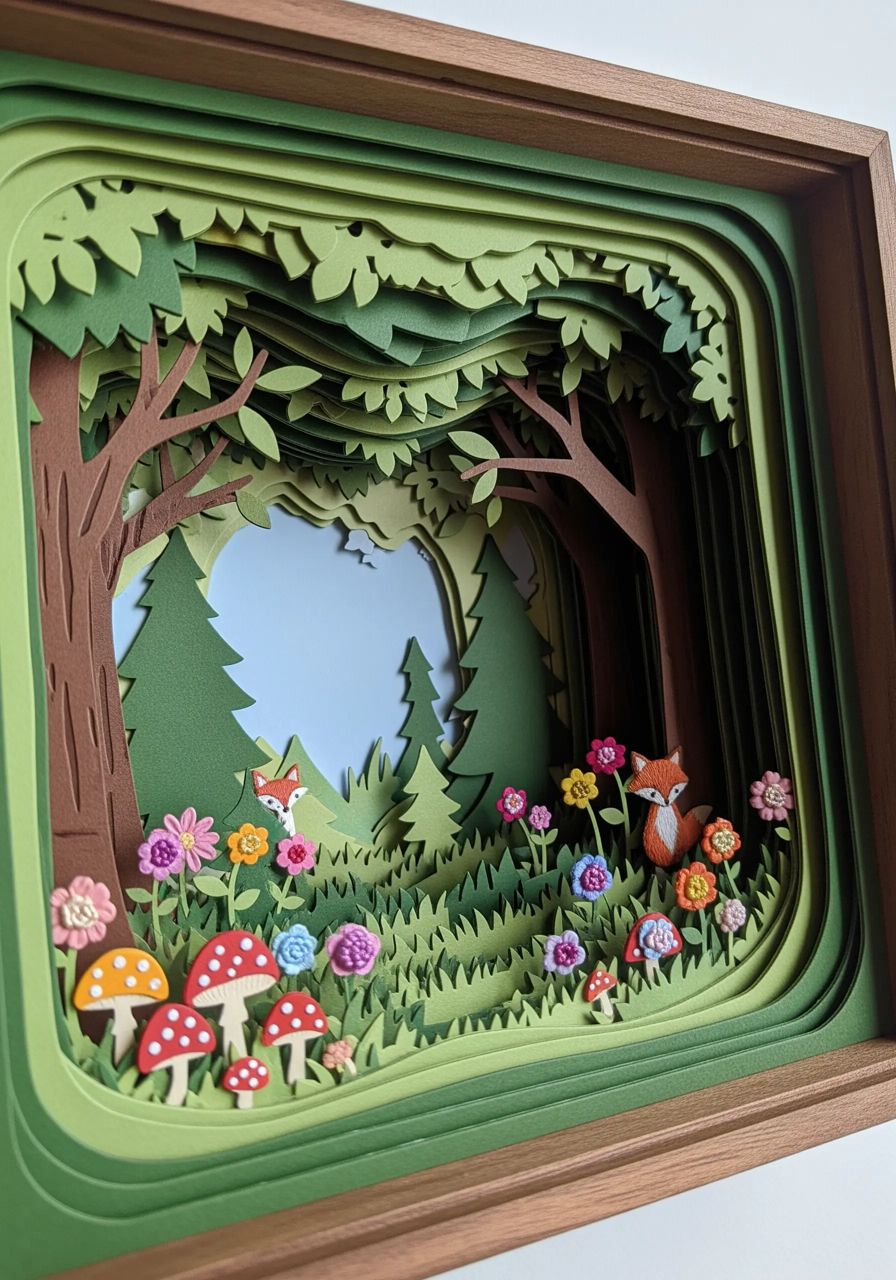 A layered papercraft diorama of a forest scene with small, embroidered fox and flower details.
