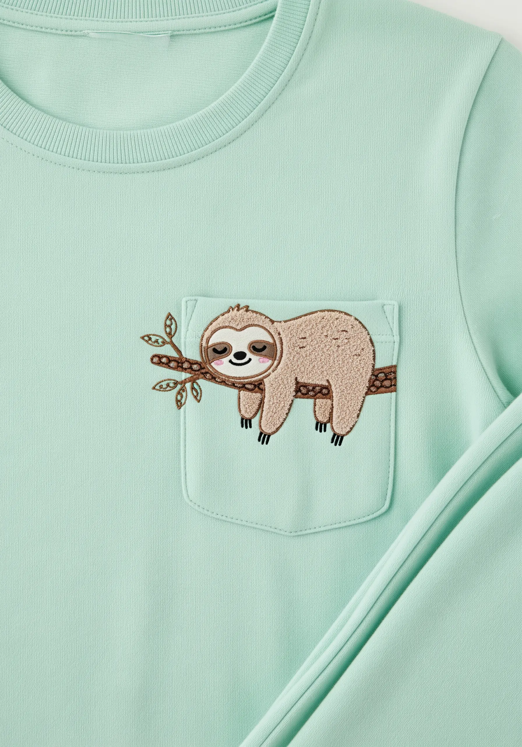 A sleeping sloth embroidered on a mint green sweatshirt, with its body on the pocket and the branch above