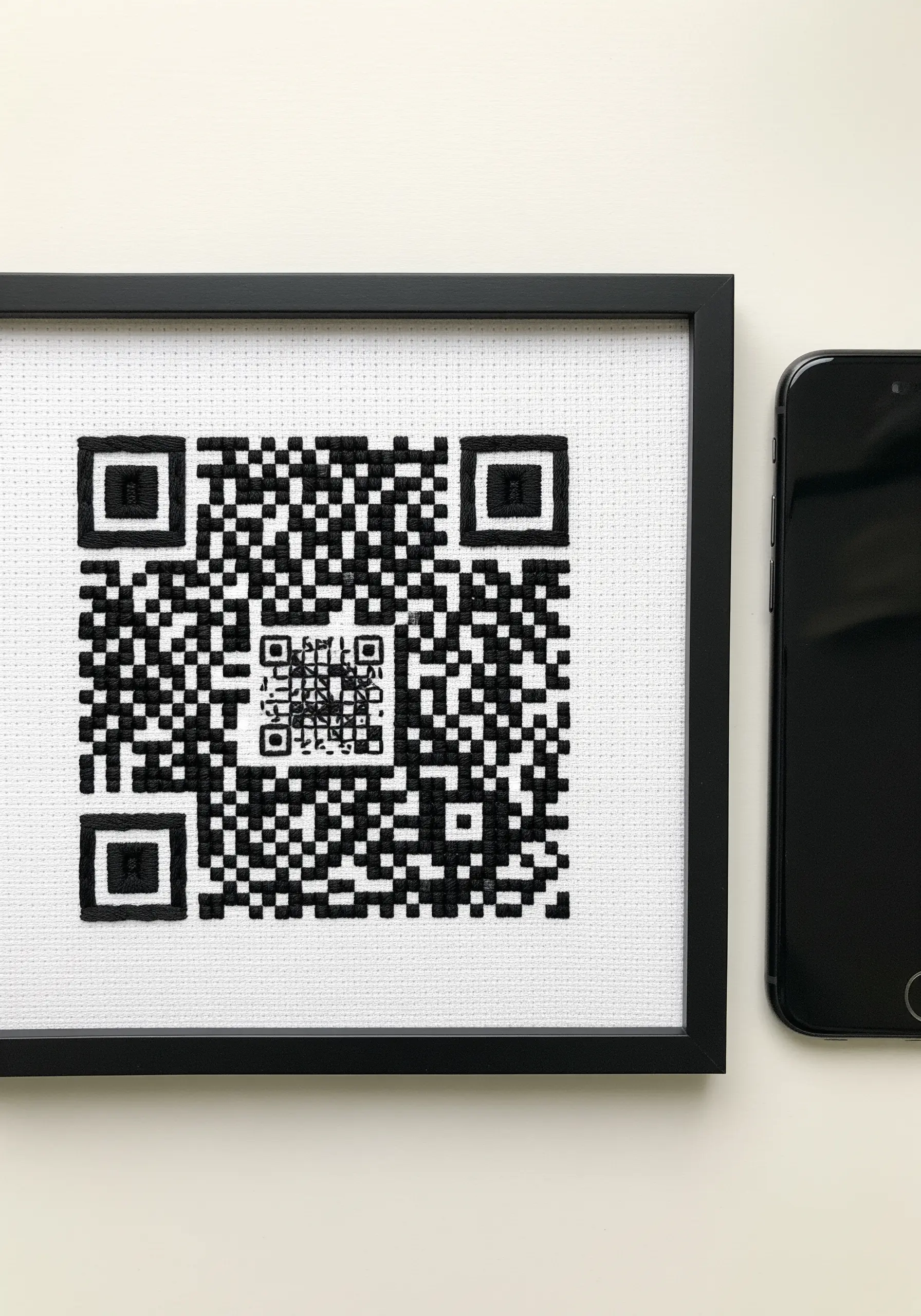 A cross-stitched QR code in a black frame, next to a smartphone.