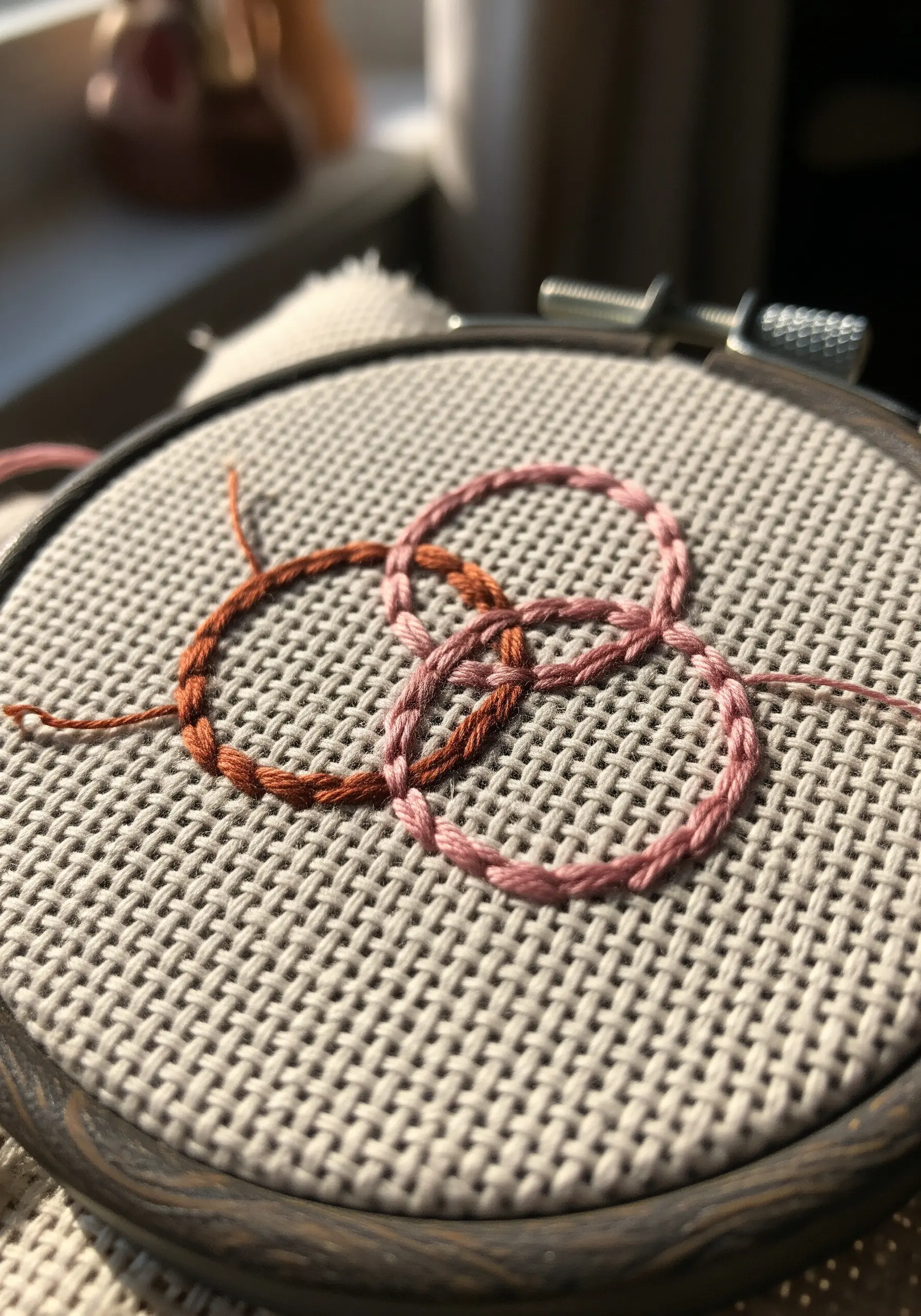 Three interlocking rings in chain stitch using variegated pink and brown thread on Aida cloth.