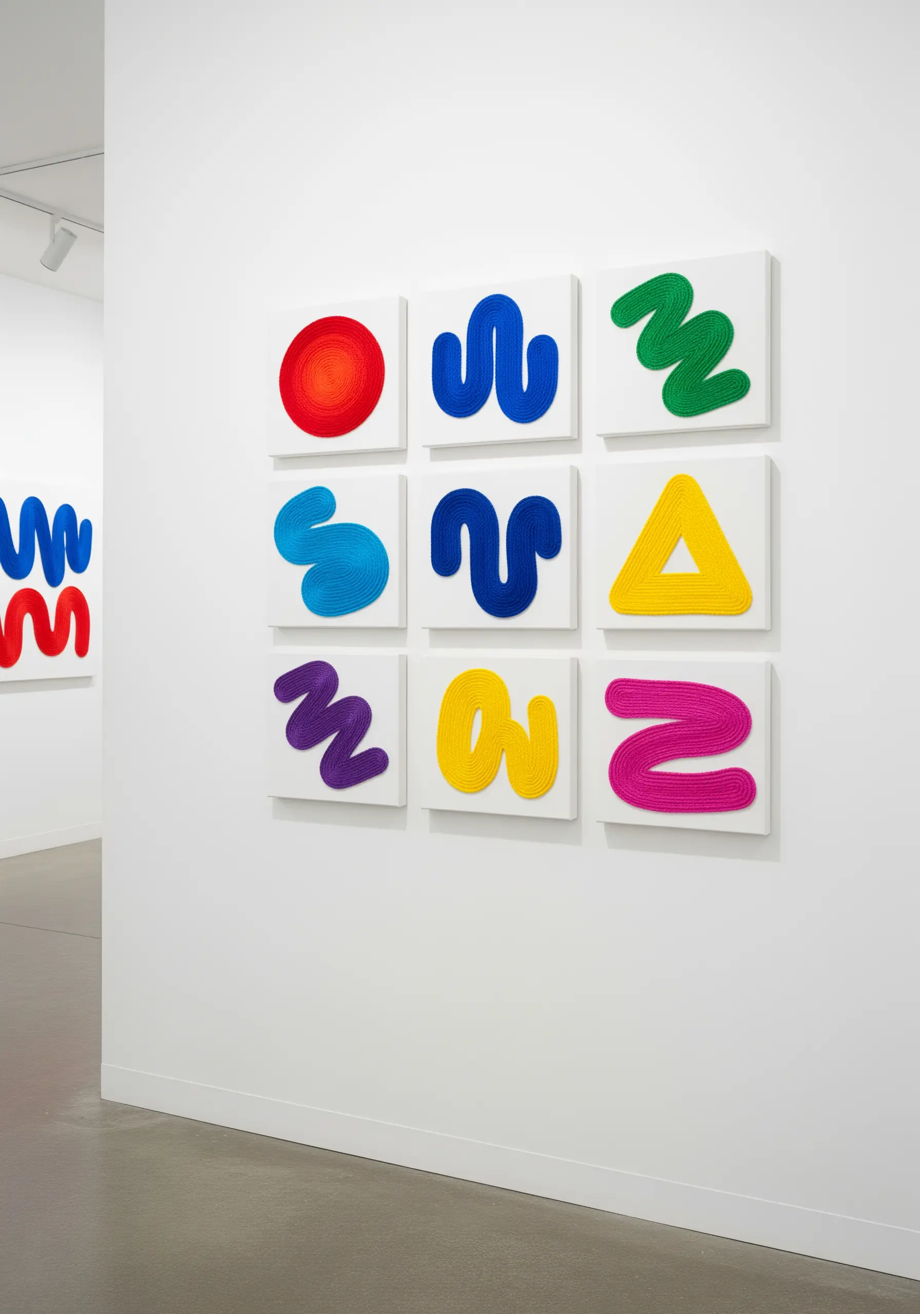 A gallery wall of nine small canvases, each featuring a colorful, abstract shape made with yarn.